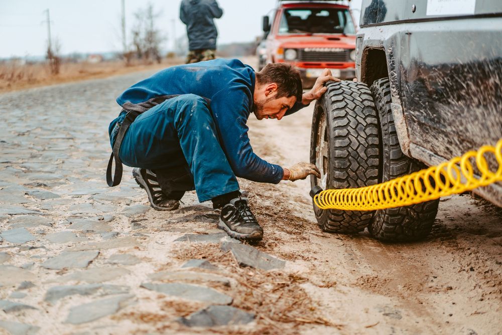 Man inflating a tire on a muddy road with a yellow hose. Red SUV in the background.