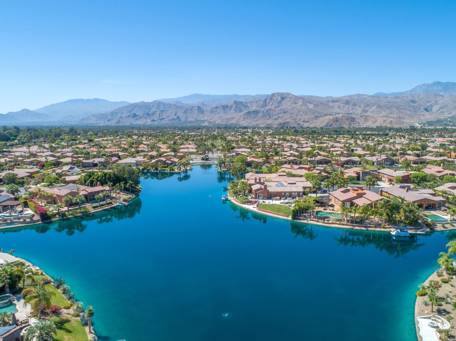 Aerial view of a blue lake surrounded by houses and palm trees, with mountains in the background under a clear sky.