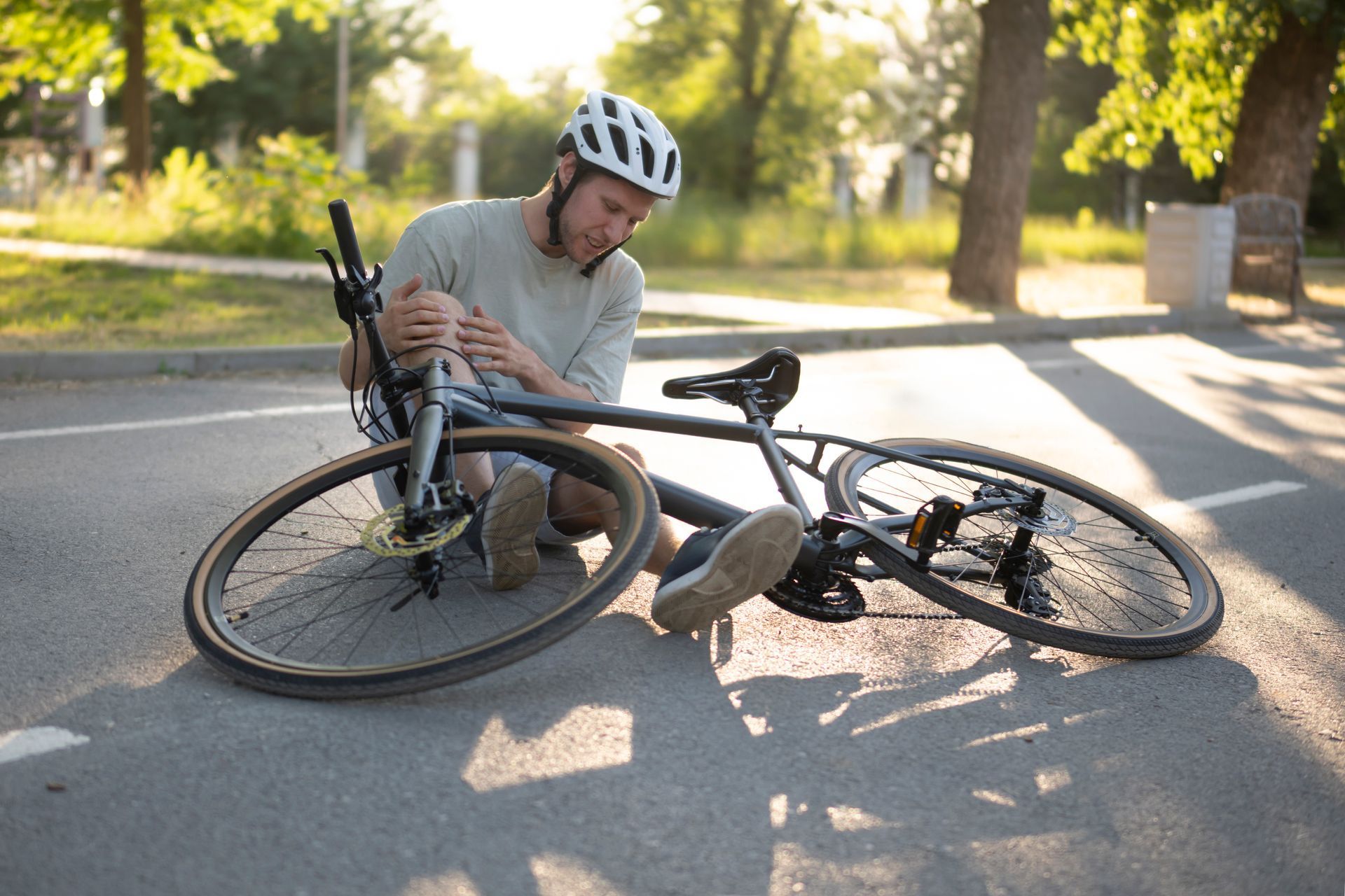 Cyclist kneeling on asphalt, bike on ground. He's holding his injured knee, wearing a helmet. Trees in background.