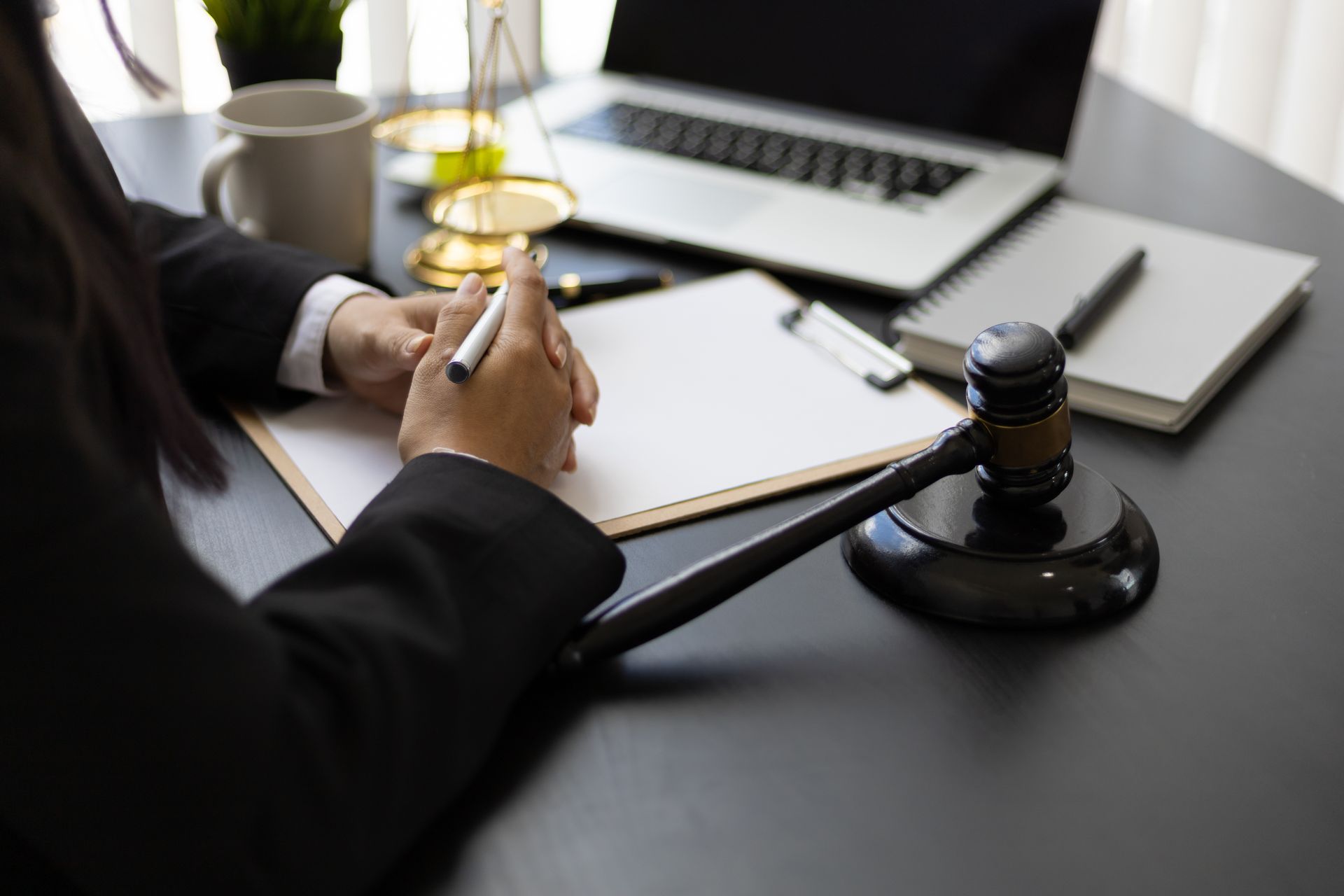 Person in a suit holds a pen over a blank document next to a gavel and laptop on a desk.