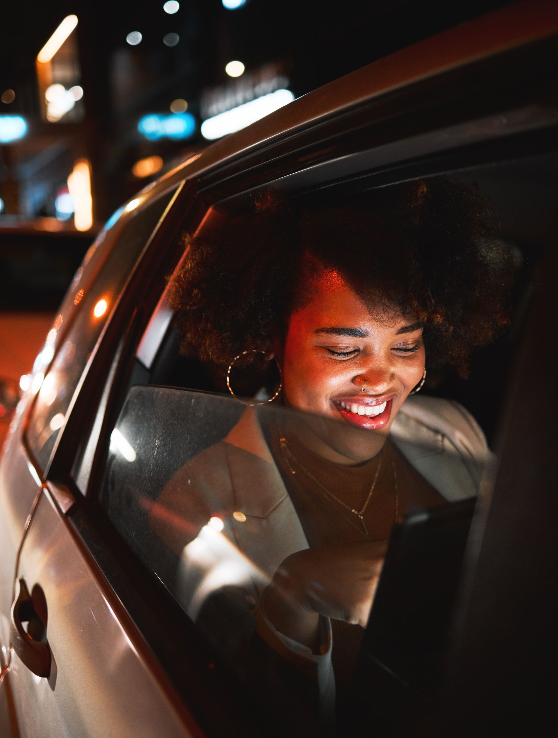 Woman in car looking at phone, smiling. Nighttime setting, window reflection.