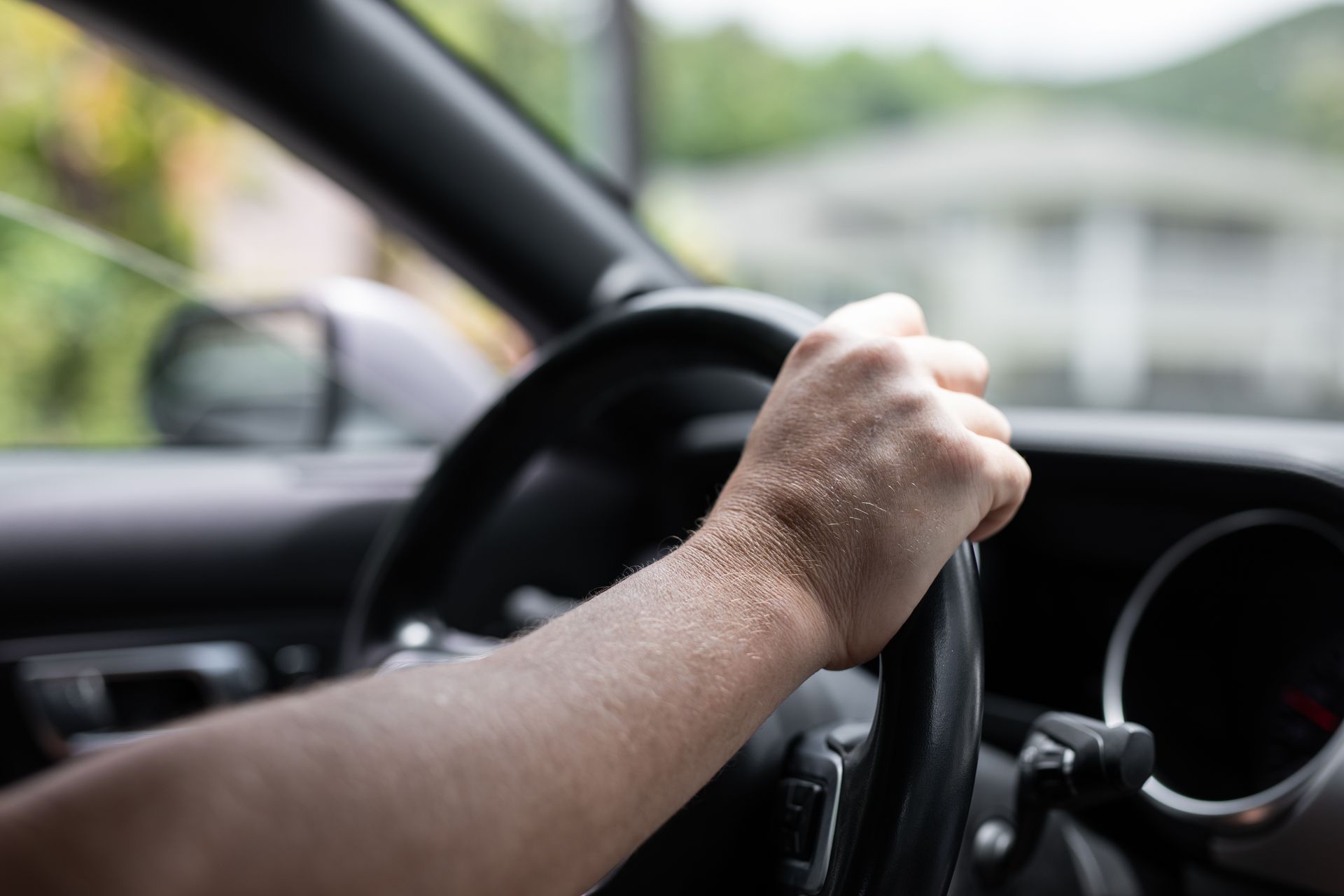 Hand gripping a car steering wheel; driving with a blurred house in the background.