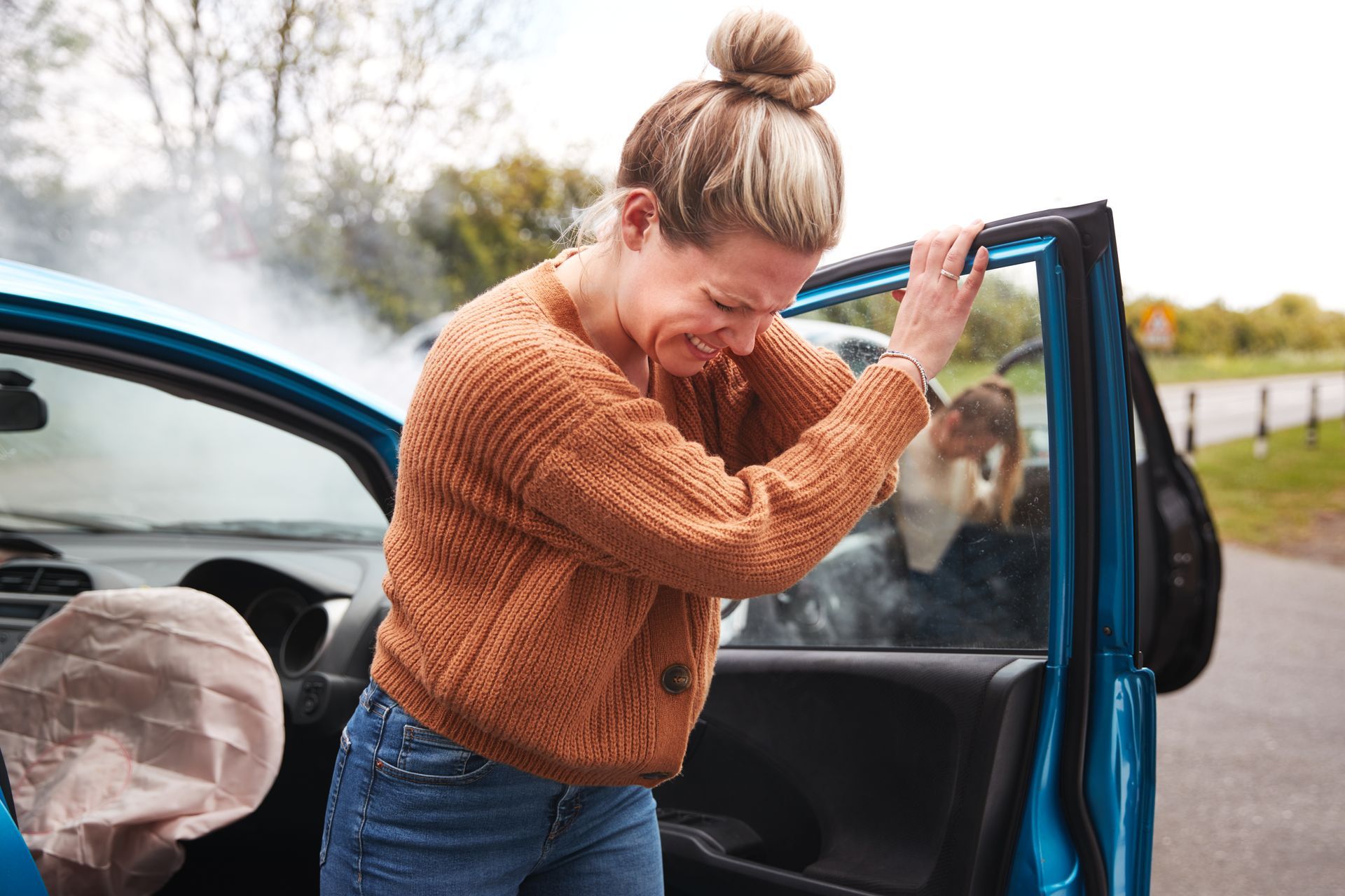 Woman exits a blue car after a crash, clutching her chest in pain, smoke rising.