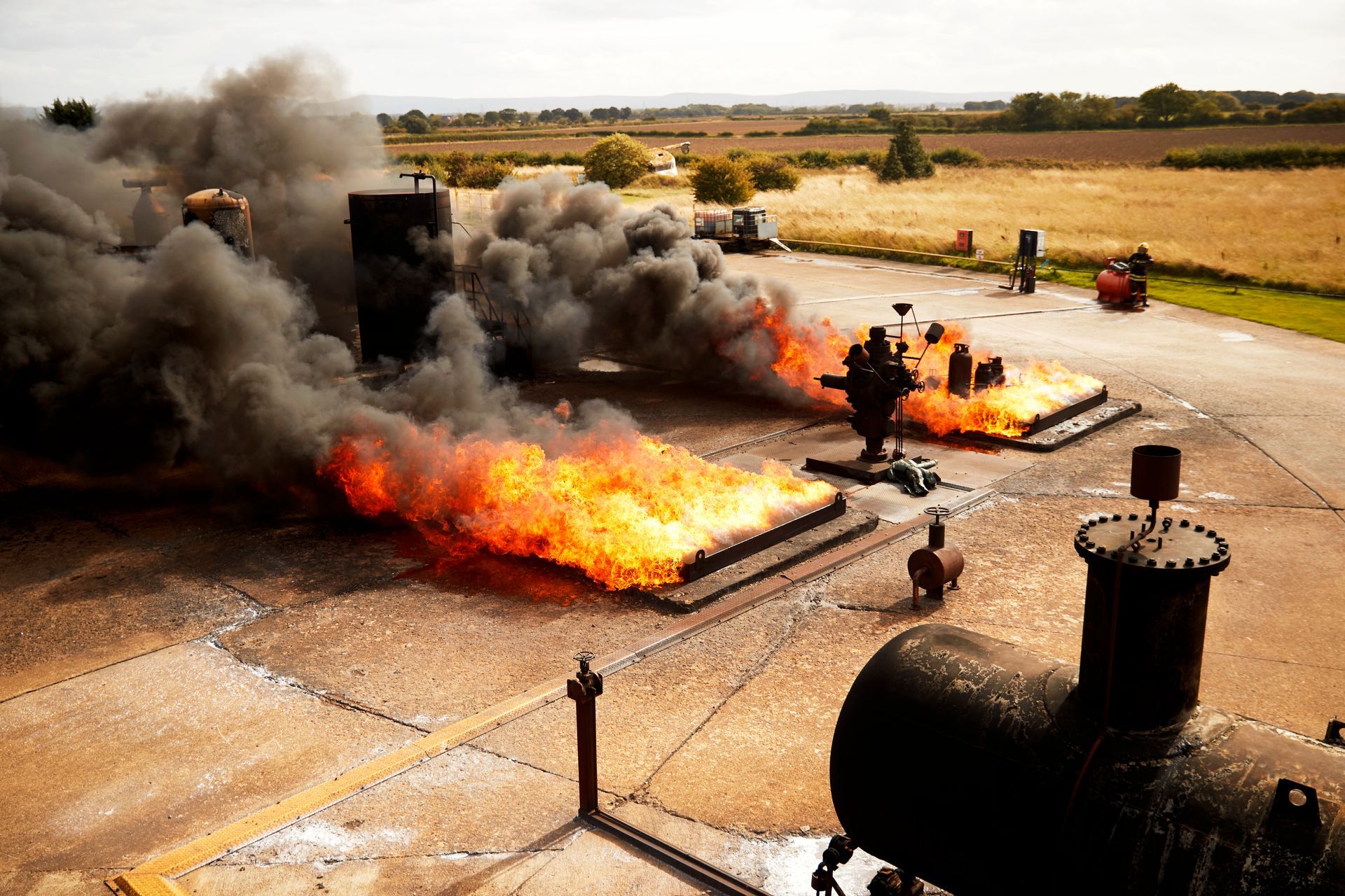 Burning structures with heavy smoke billowing into the sky on a concrete platform in an open field.