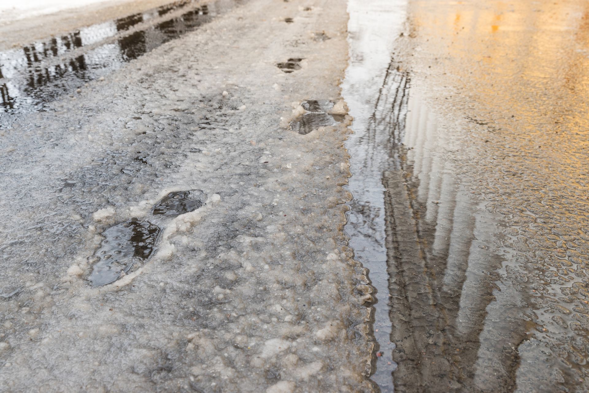 Puddle on a concrete surface reflects a building's columns; muddy footprints visible.