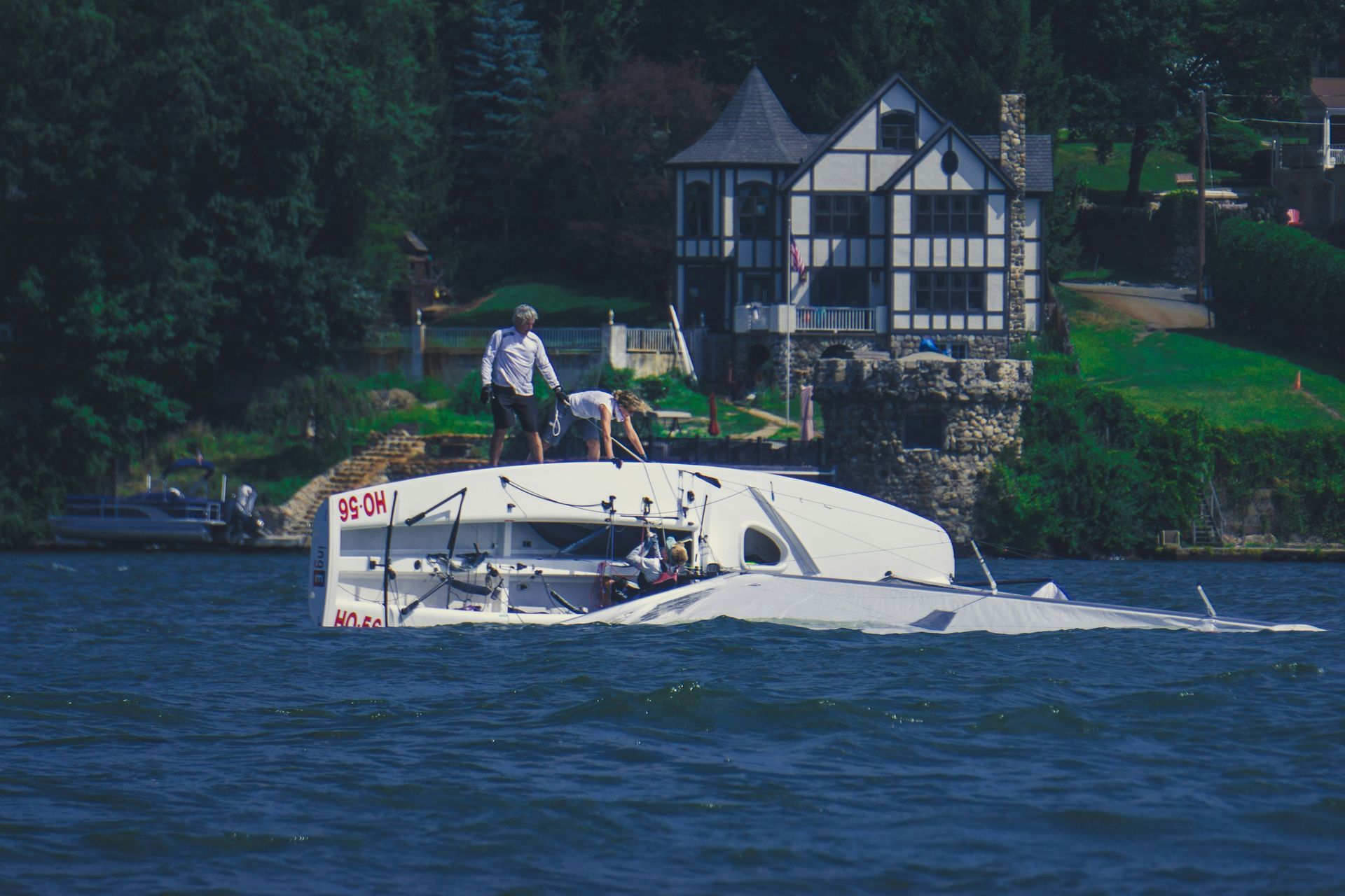 A partially submerged white sailboat with people on top near a Tudor-style house by the water.