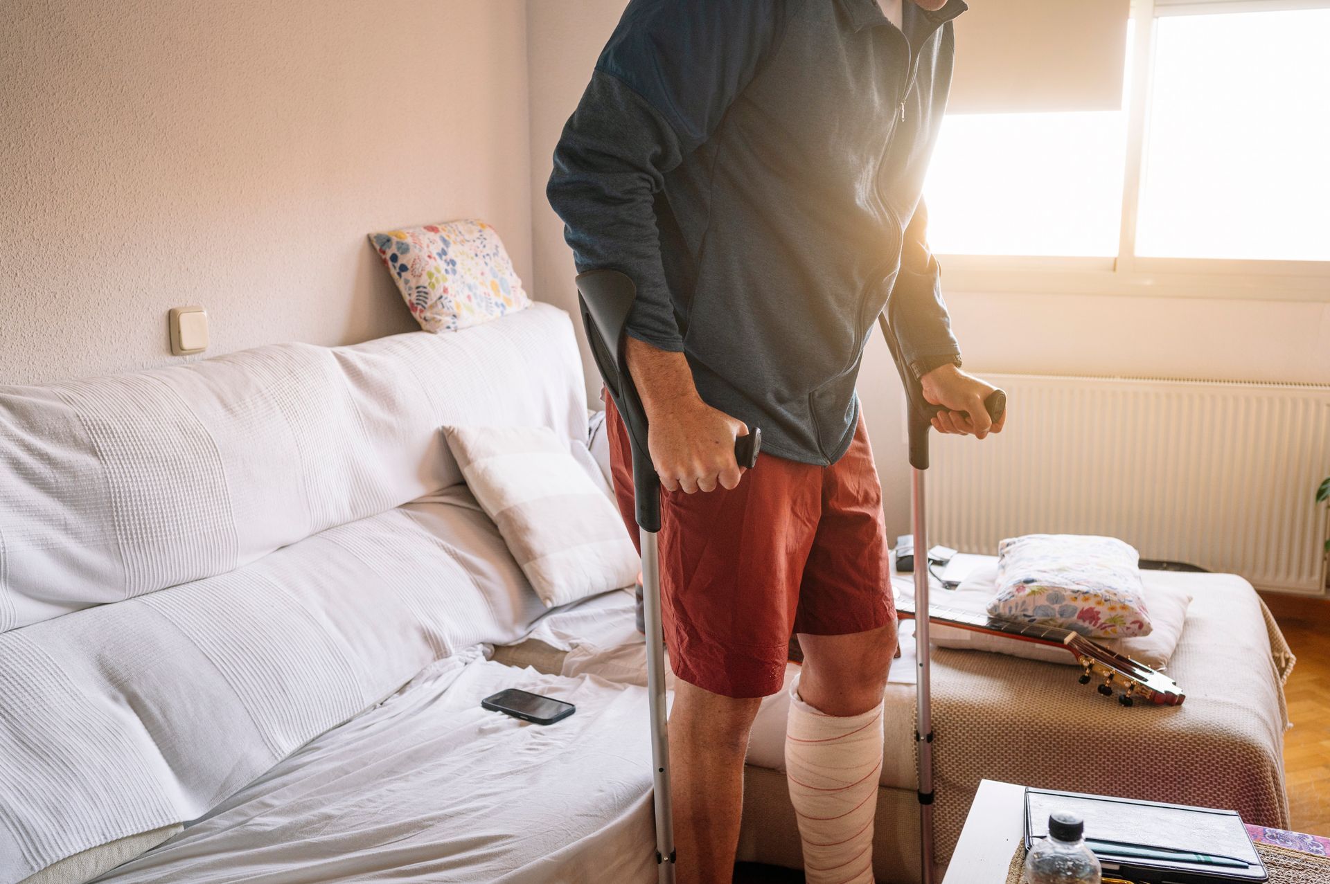 Man with leg cast using crutches indoors near a couch and window.