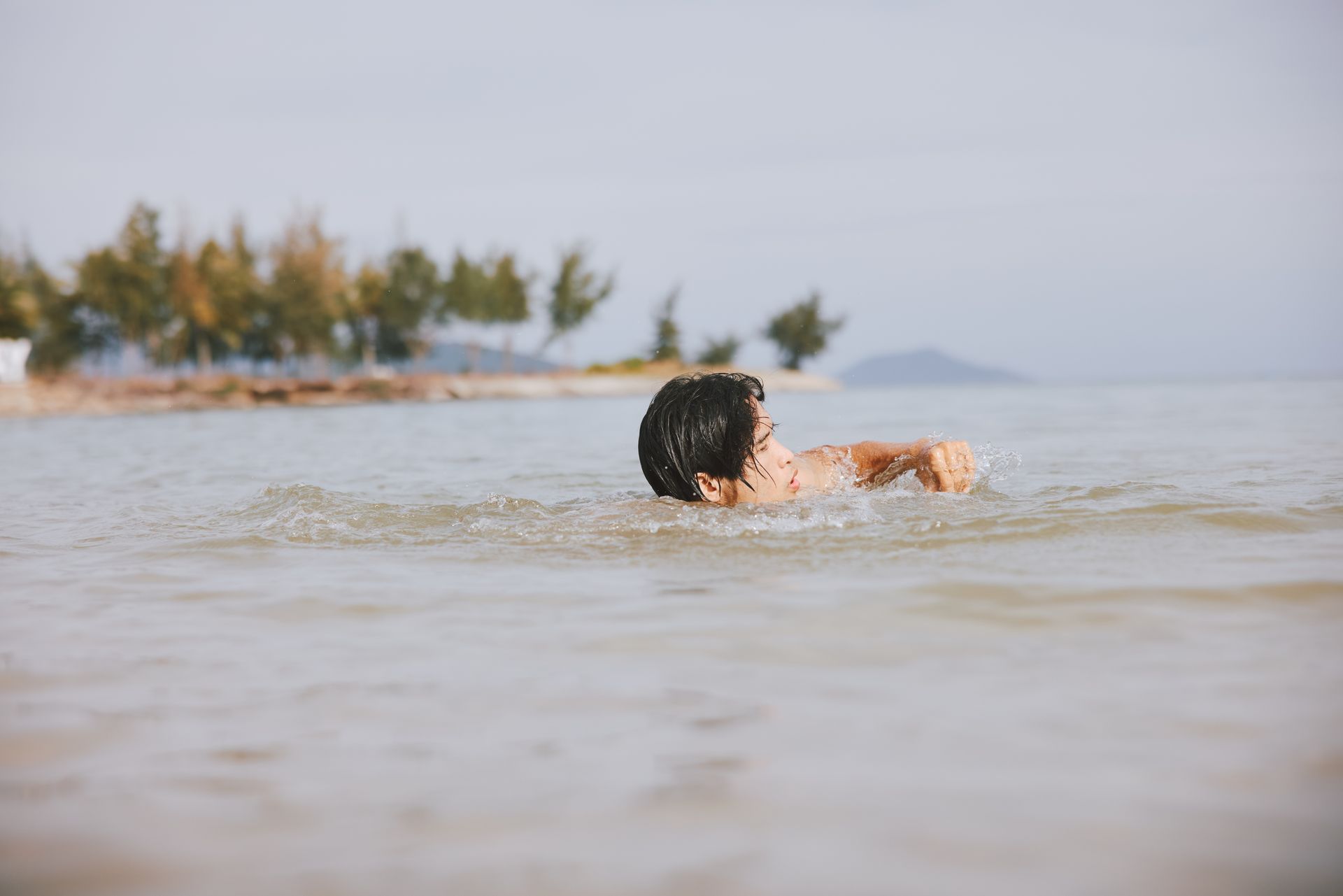 Person swimming in shallow water near a shoreline with trees, ocean, and overcast sky.