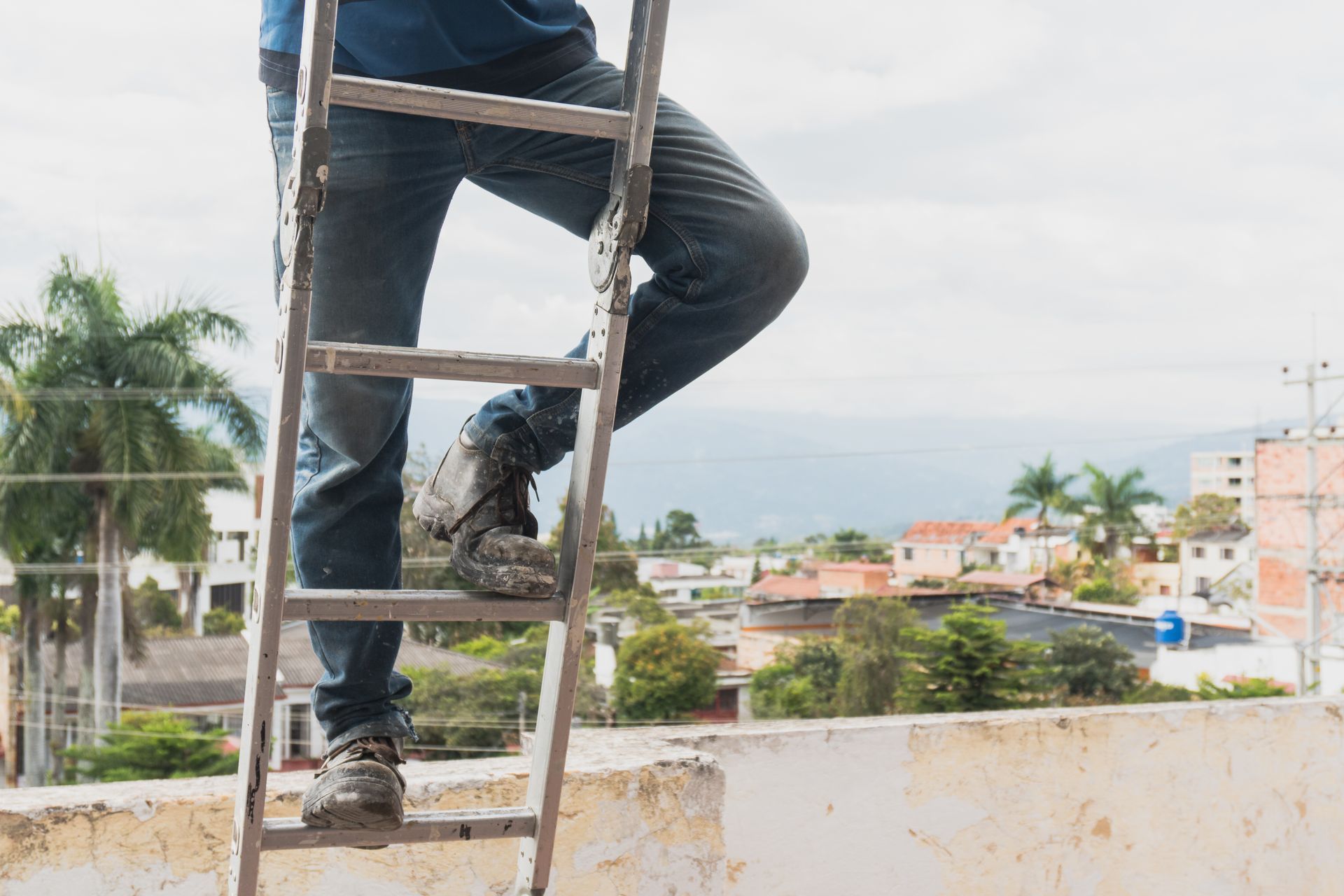 Person climbing a metal ladder onto a rooftop, city buildings and sky in background.