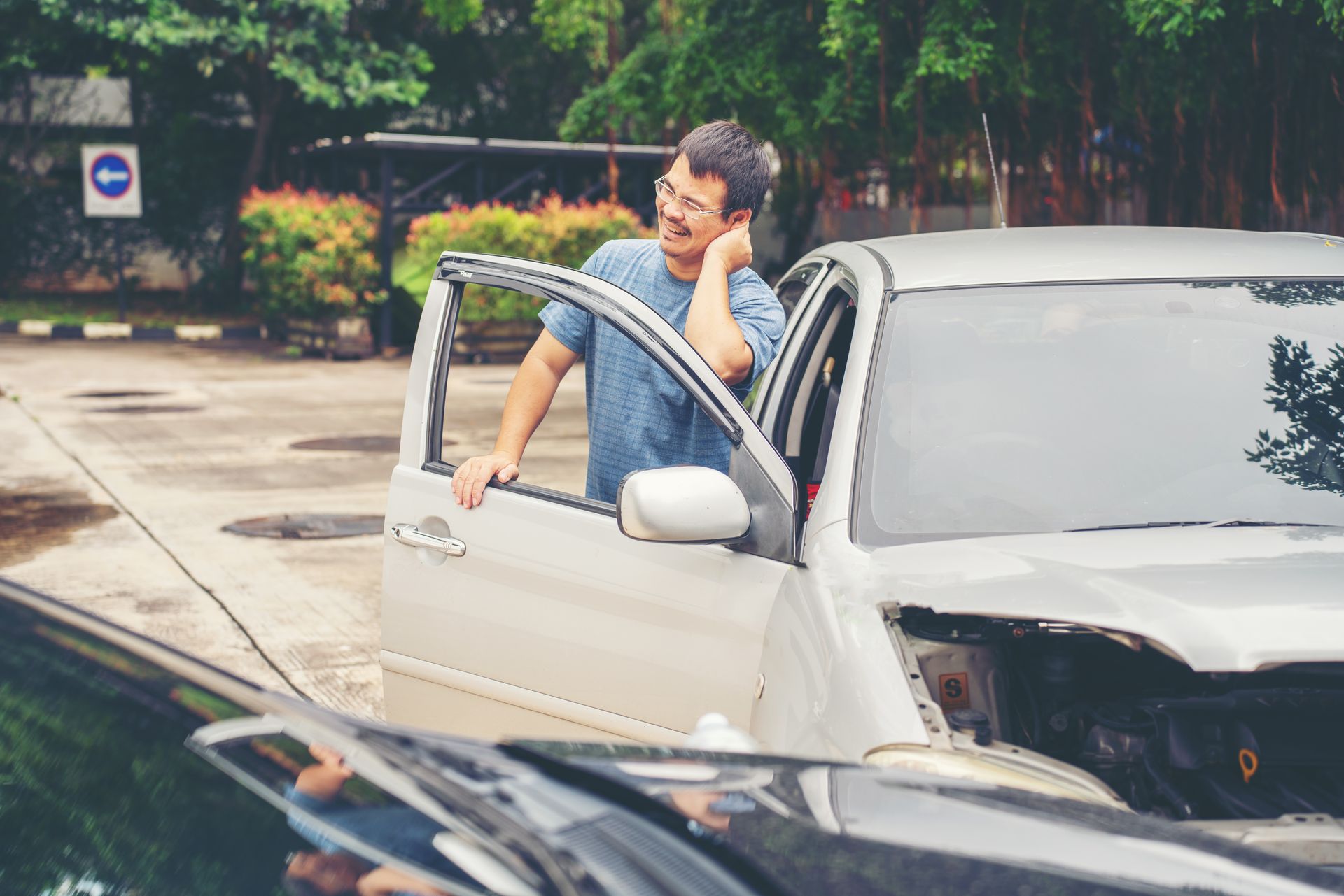 Man with glasses holds his neck, standing by damaged car after accident.