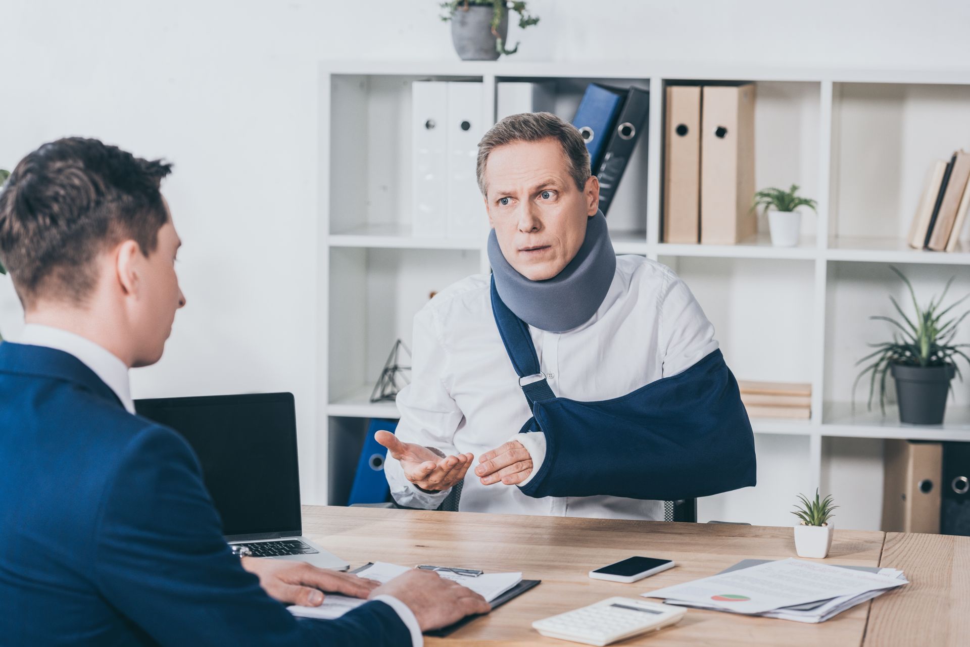 Man with arm sling and neck brace talking to another man at an office desk.