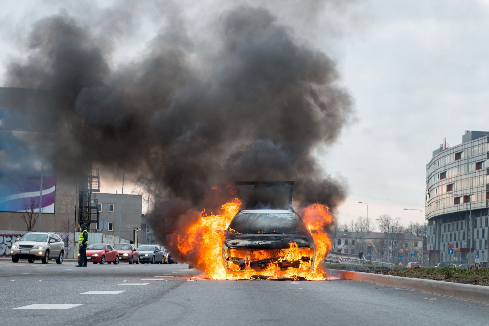 Burning car on a city street, flames and thick black smoke billowing. Other vehicles and a building are visible.