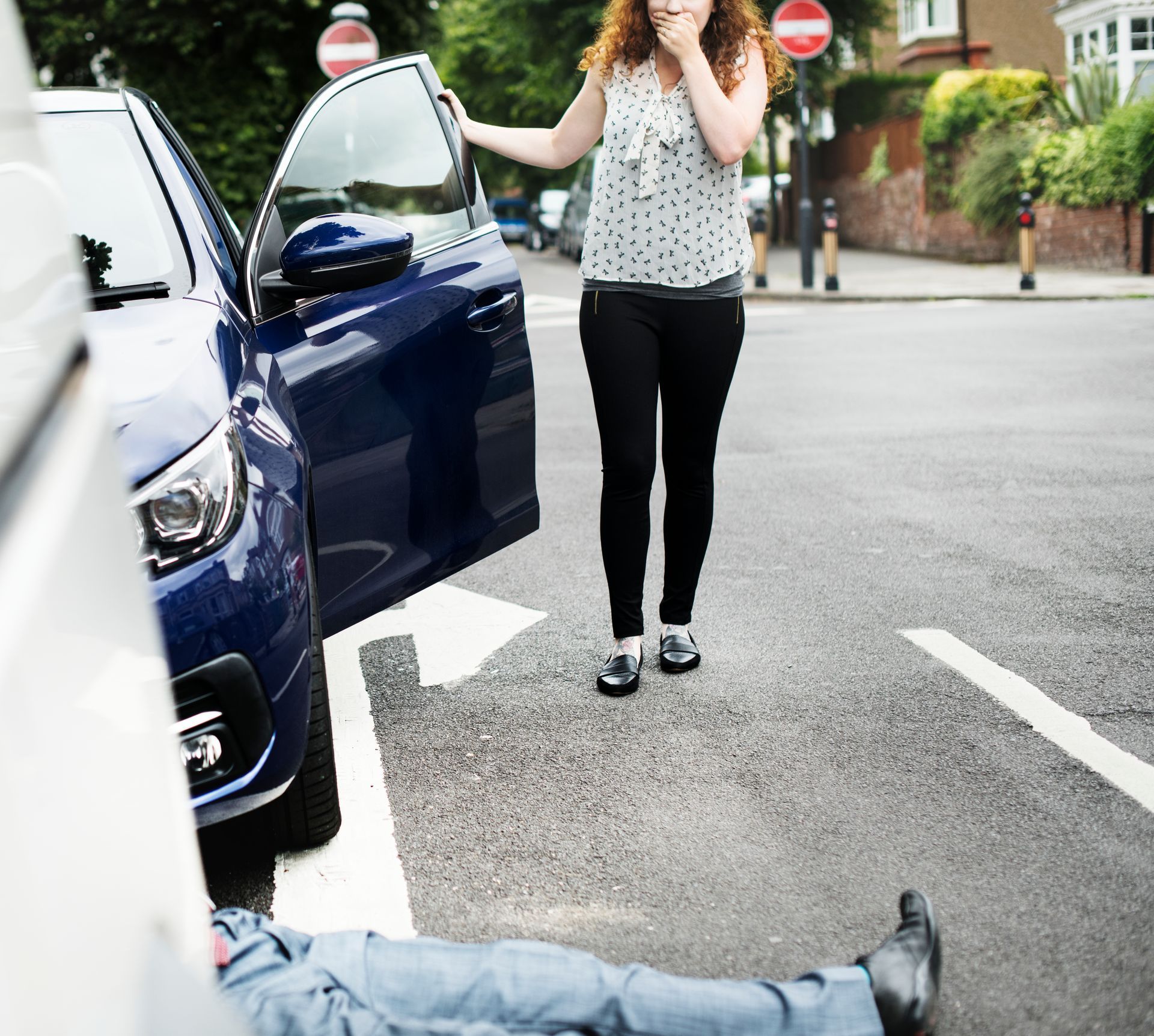 Woman near blue car, looking at person lying on the street after an accident.