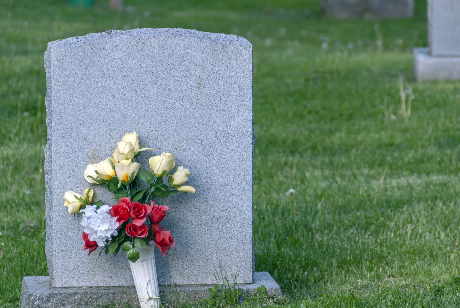Blank gray headstone with bouquet of red, white, and yellow flowers in a grassy cemetery.