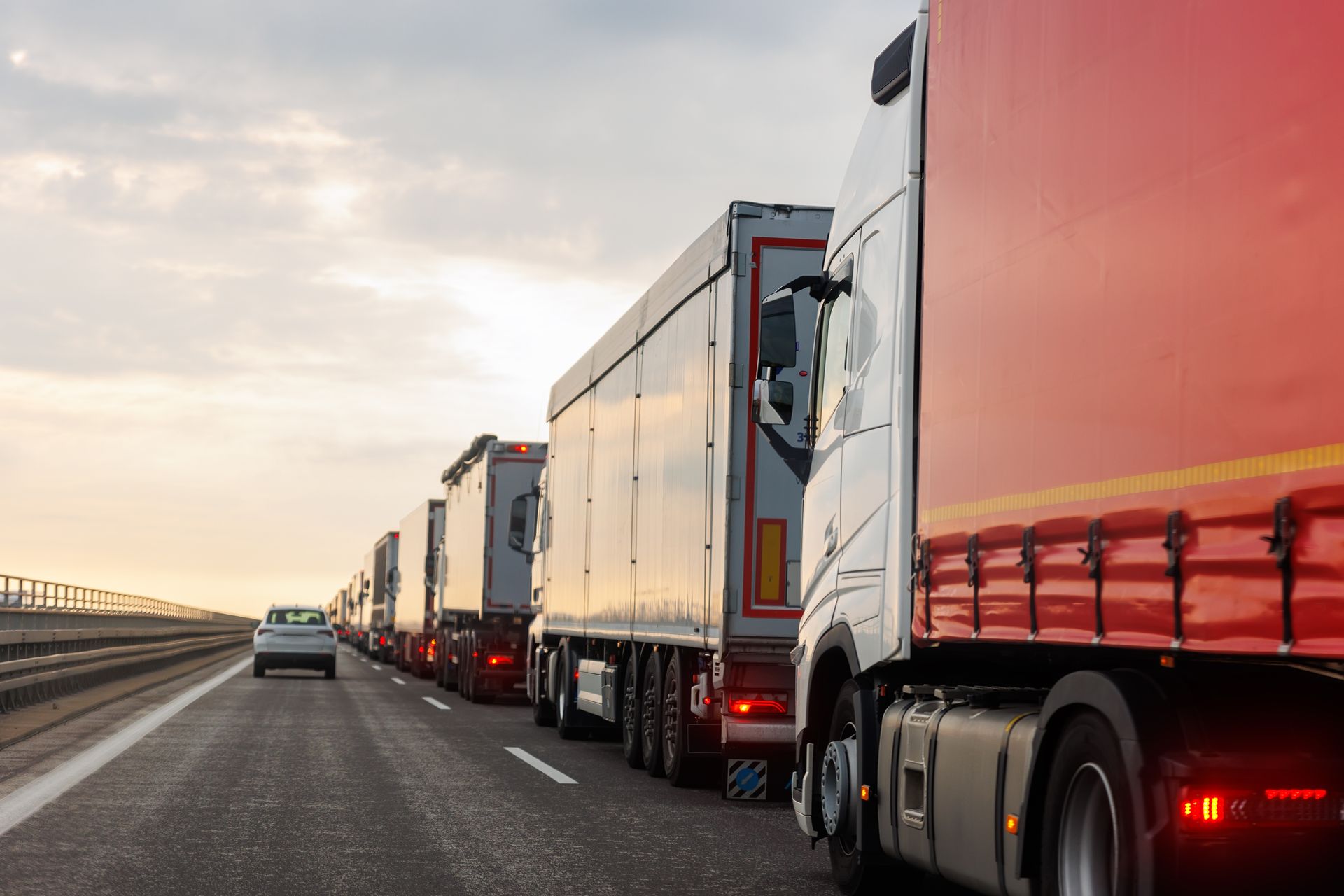 Line of semi-trucks on a highway, with a car in the left lane; overcast sky.