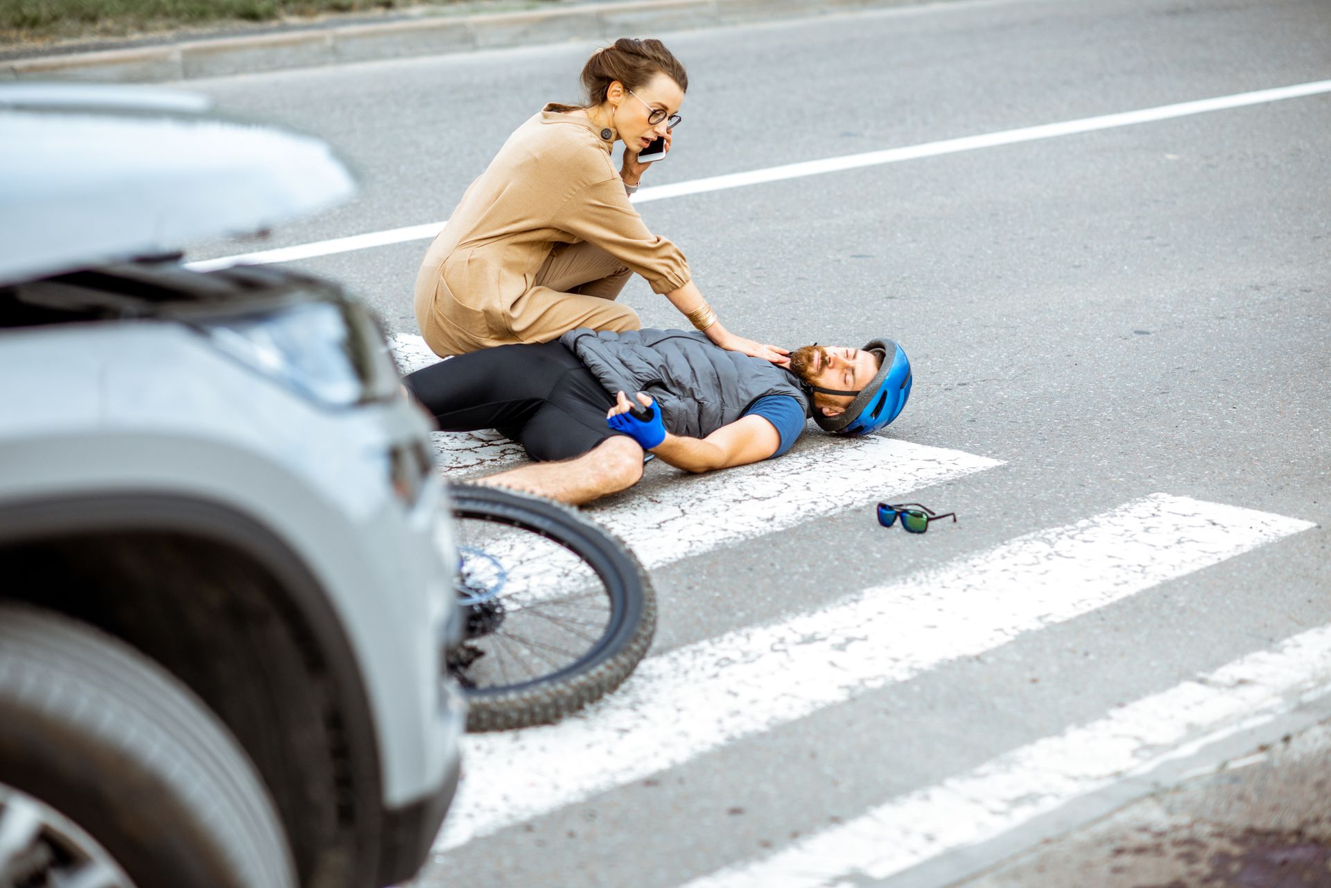 Woman on phone kneels beside cyclist lying on a crosswalk after a car accident; bicycle and car in scene.