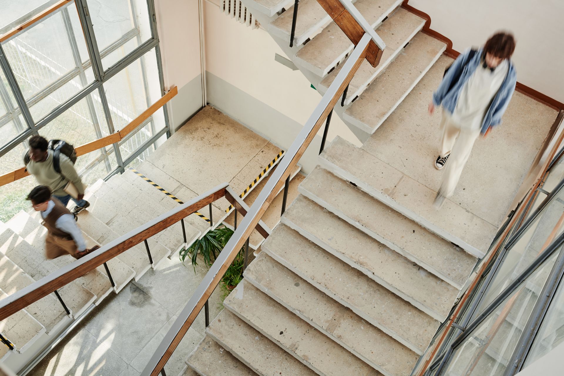 People on a staircase in a building. One person descends, two others ascend. Beige and brown tones.