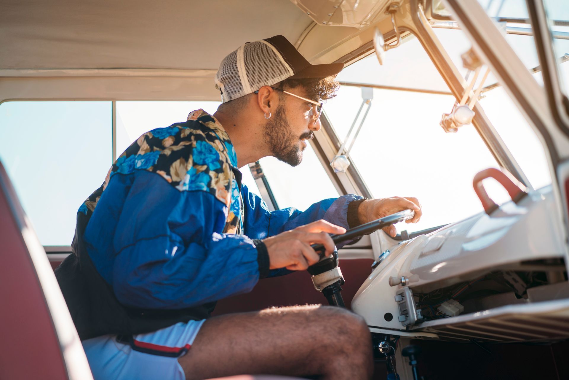 Man driving a vintage vehicle, wearing a blue jacket and a trucker hat. Bright sunlight through the windshield.