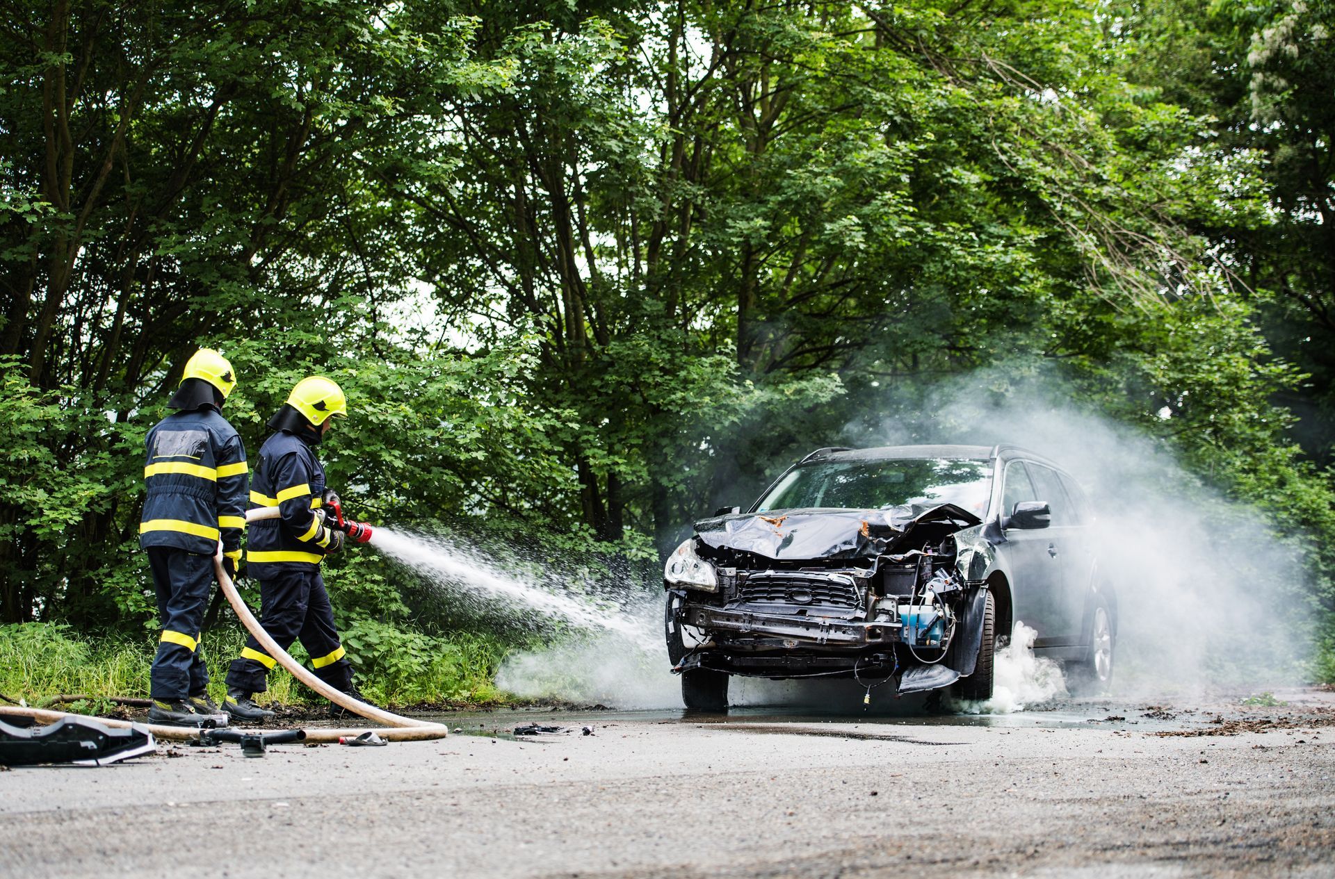 Firefighters extinguish a damaged car on a road, smoke rising, with trees in the background.