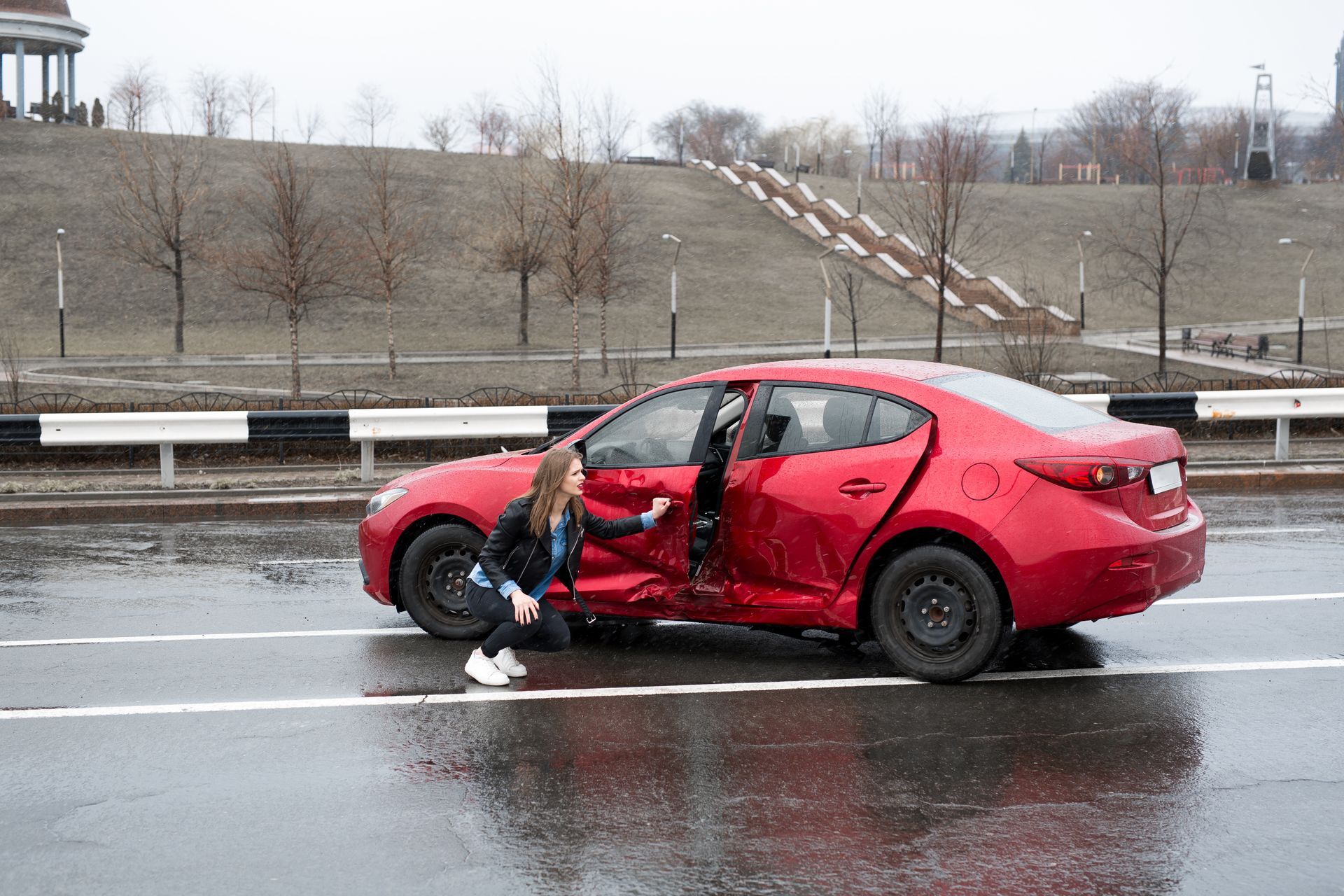 Woman examining red car damaged on the side of a wet road. A park is in the background.