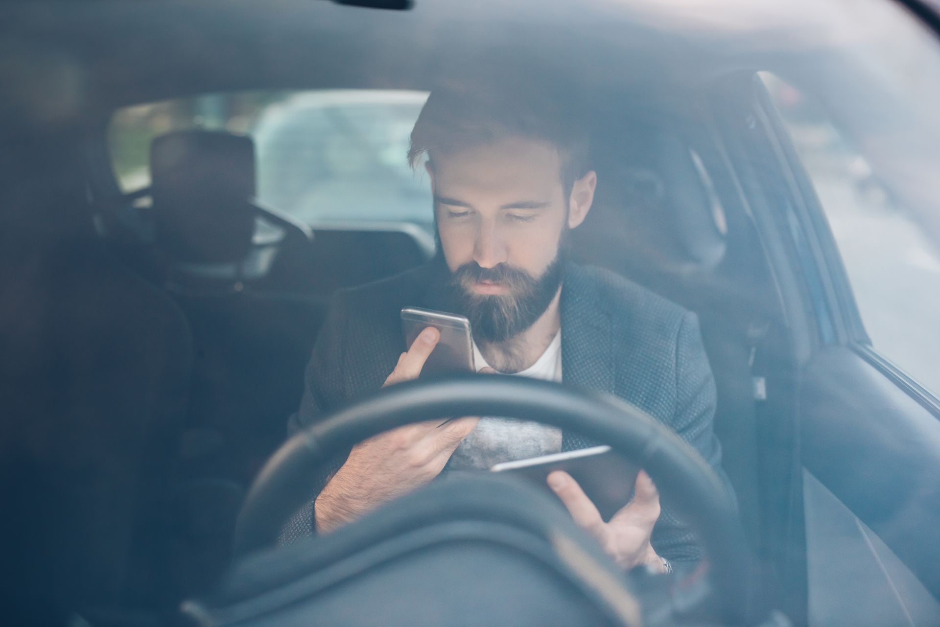 Man in a car, looking down at a phone and tablet simultaneously. Steering wheel is visible.