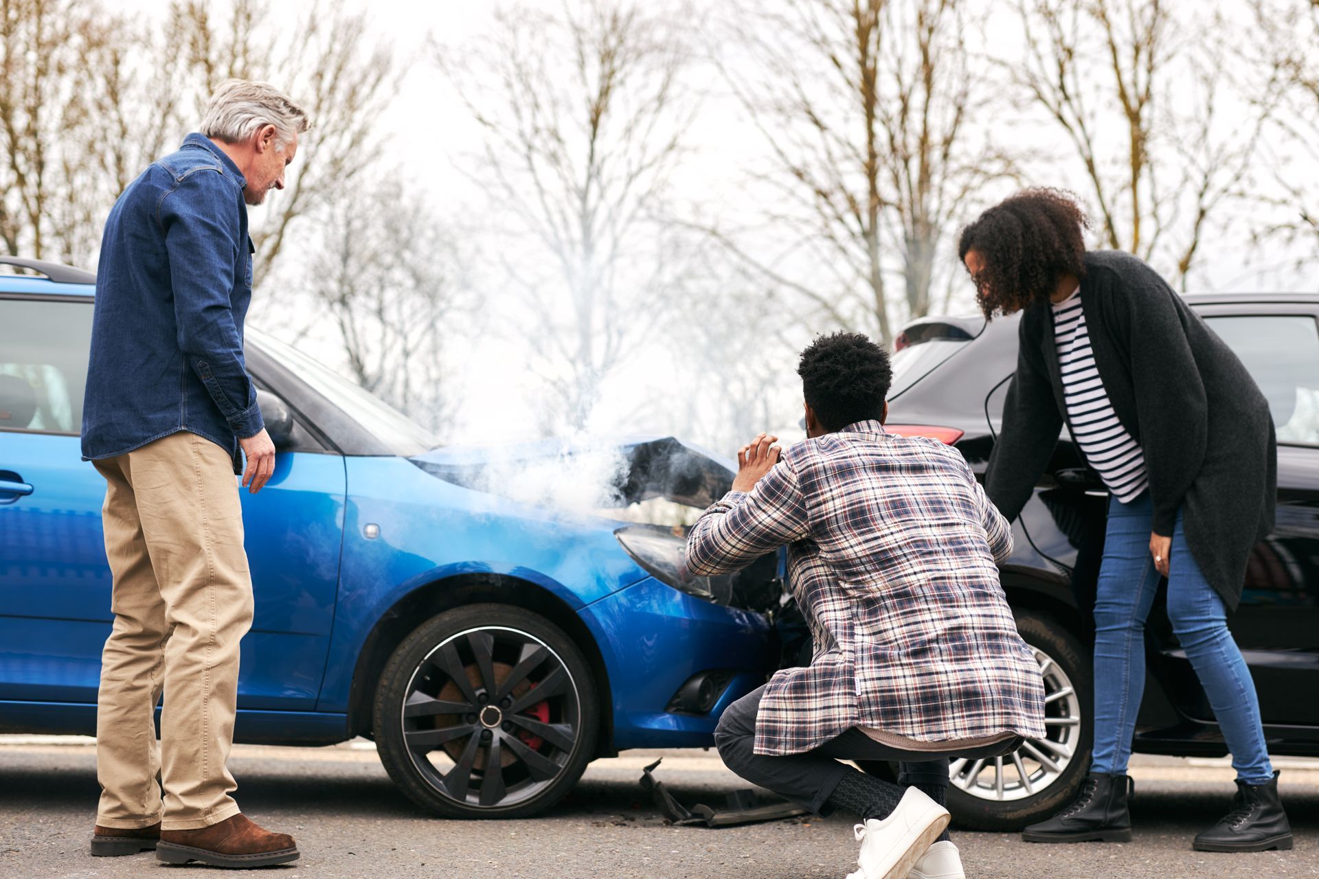 People inspecting a damaged, smoking blue car after a collision on a street.