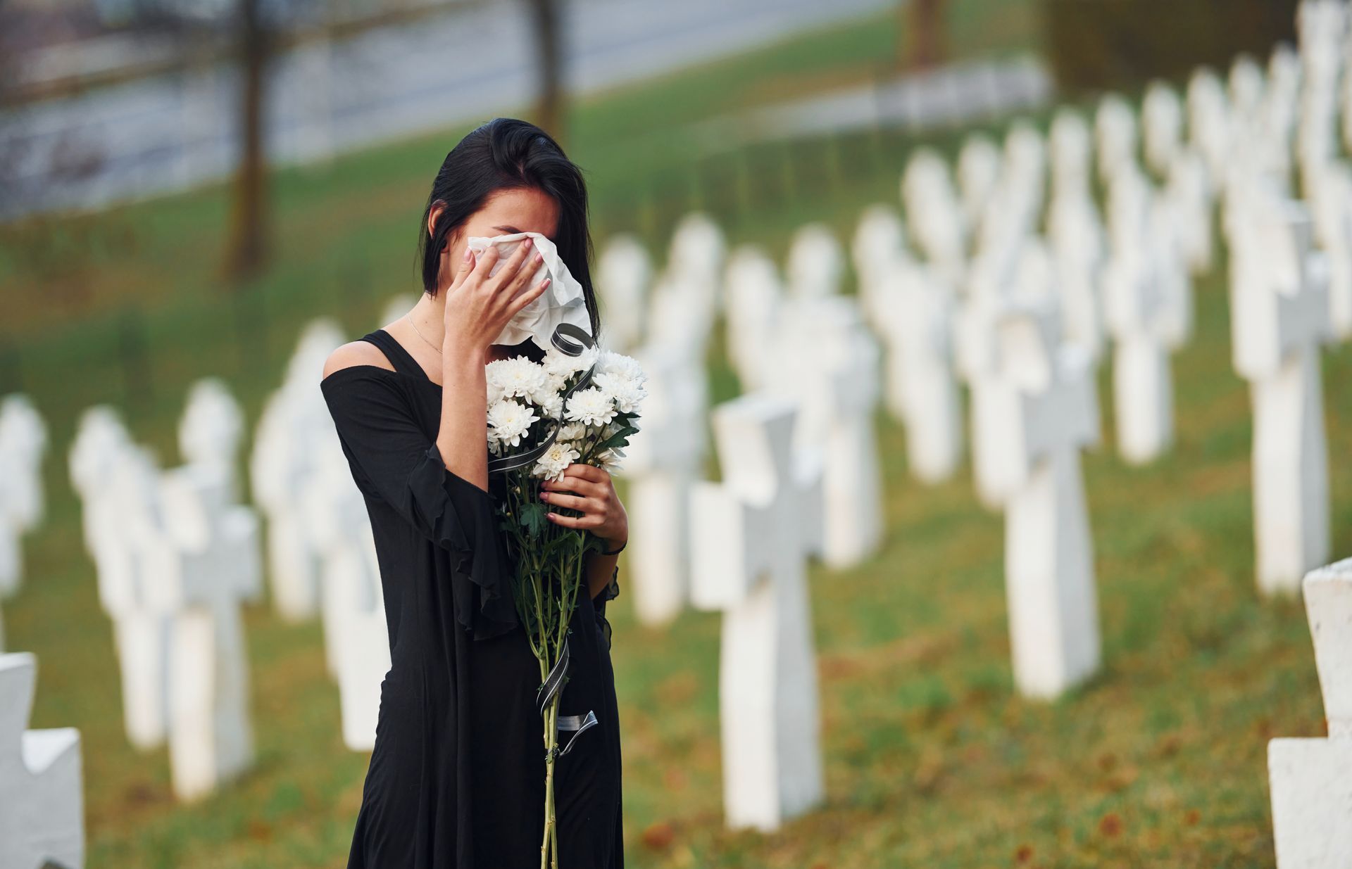 Woman in black, weeping at a gravesite, holding flowers. White crosses in the background.
