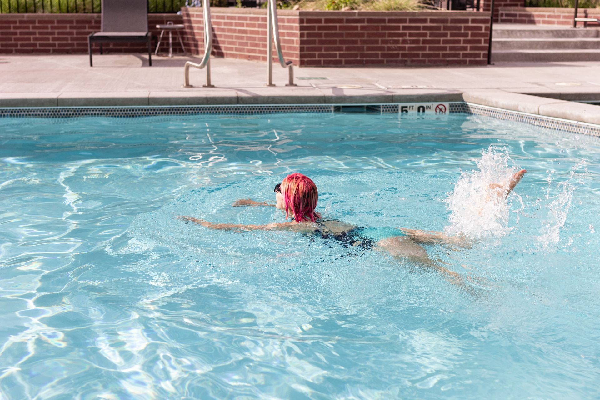 Person swimming in a pool, kicking up a splash. Bright blue water, sunny outdoor setting.