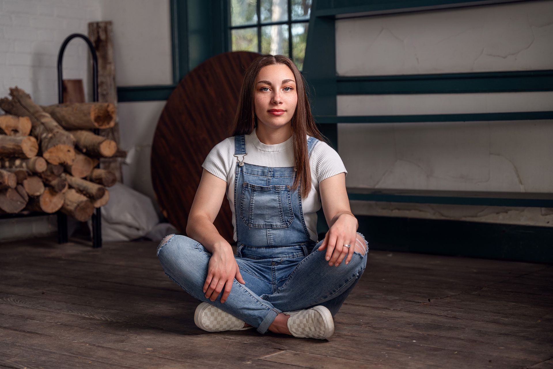 A woman in overalls is sitting on the floor with her legs crossed.