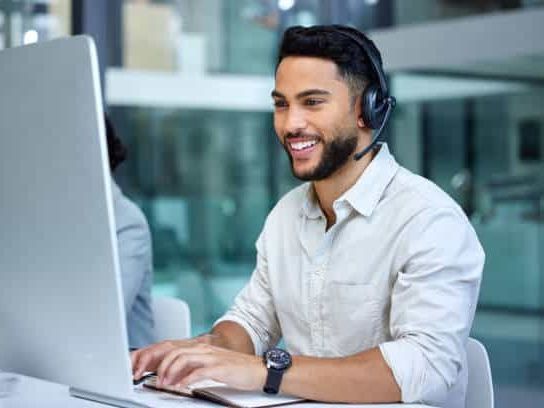 A man is sitting at a desk with a laptop and talking on a cell phone.