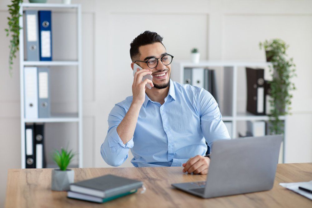 A man is sitting at a desk with a laptop and talking on a cell phone.