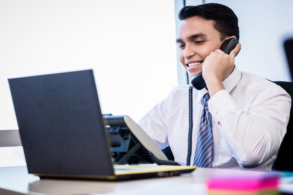 A man is sitting at a desk with a laptop and talking on a cell phone.