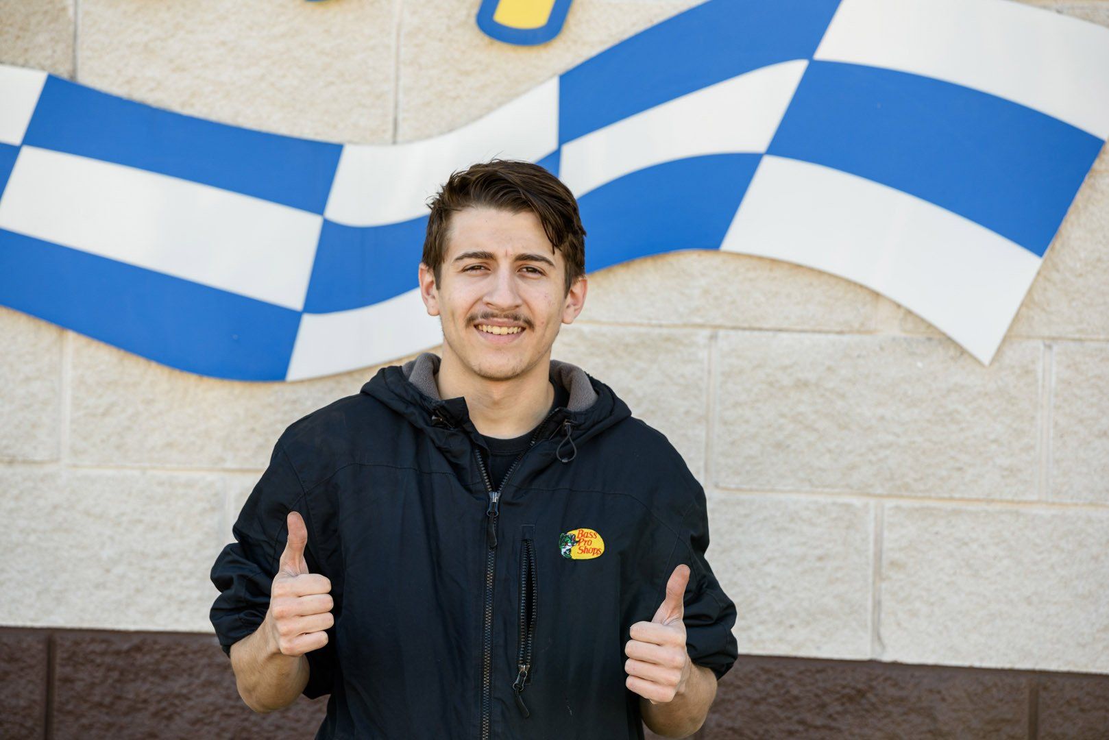 Man in black jacket giving thumbs-up, smiling in front of a blue and white checkered flag.