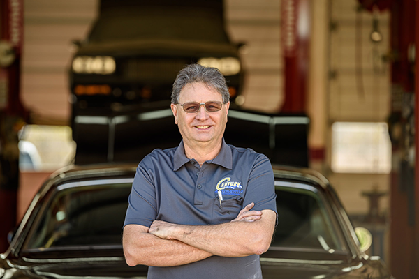 Man with arms crossed, smiling in front of a car at a mechanic shop, wearing a blue shirt and glasses.