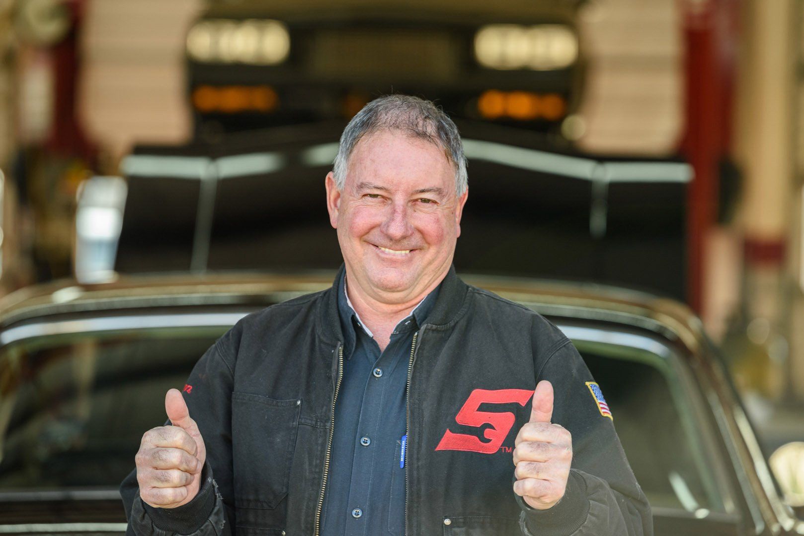 Mechanic in black uniform gives thumbs up in front of a classic car, shop setting.