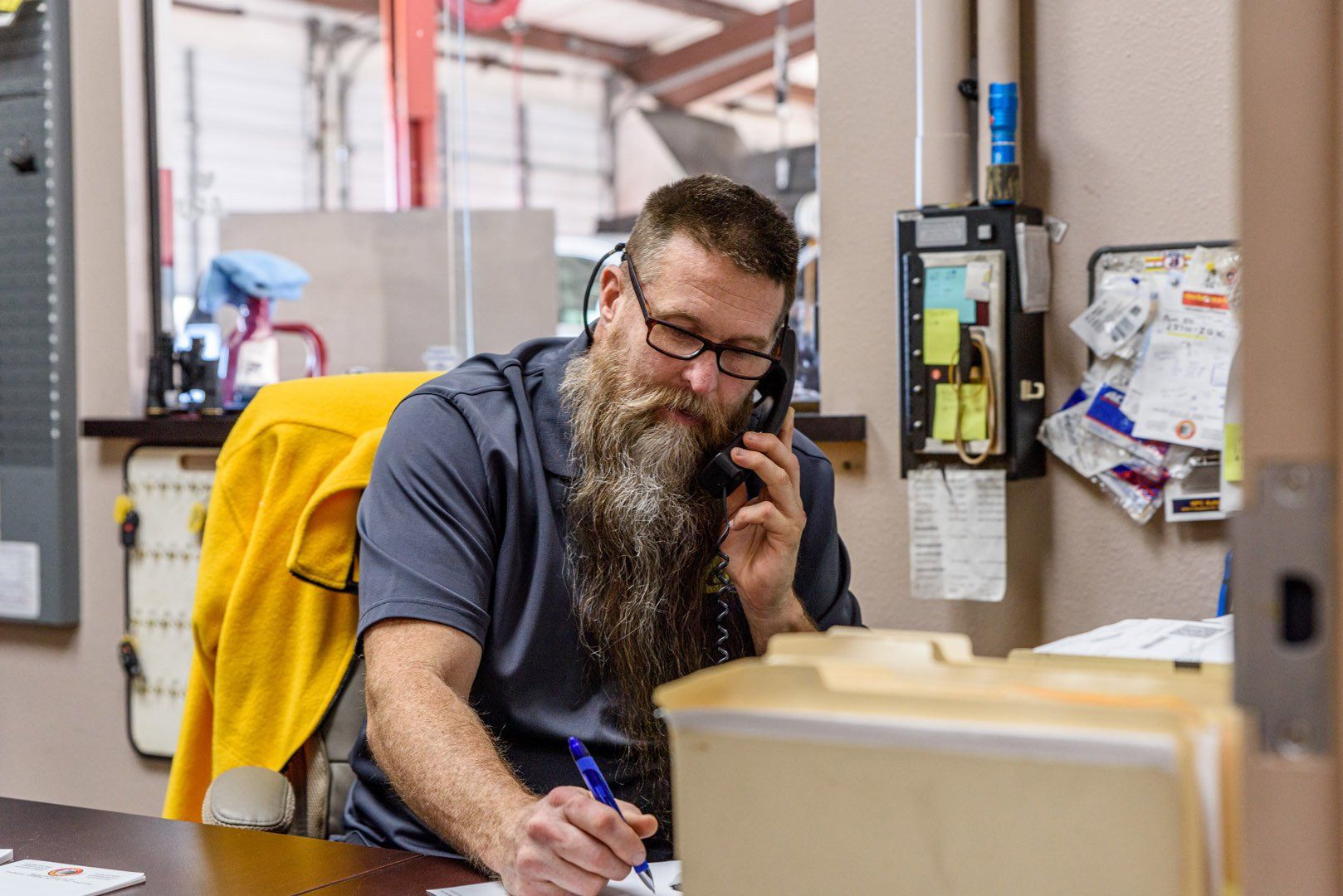 Man with beard on phone, writing at desk in an office. Yellow jacket hanging, files visible.