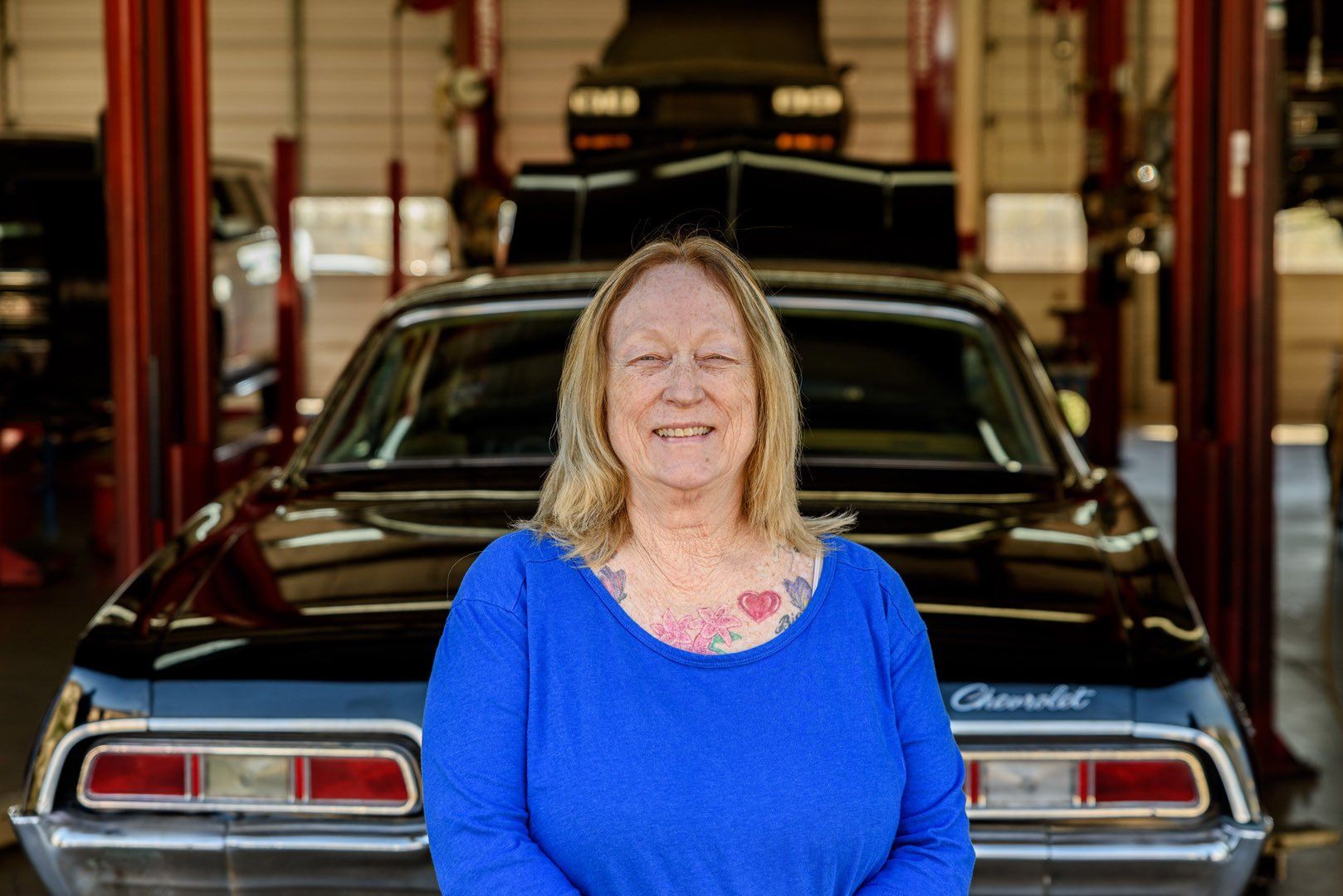 Woman in blue shirt smiles in front of a black classic car at an auto shop.