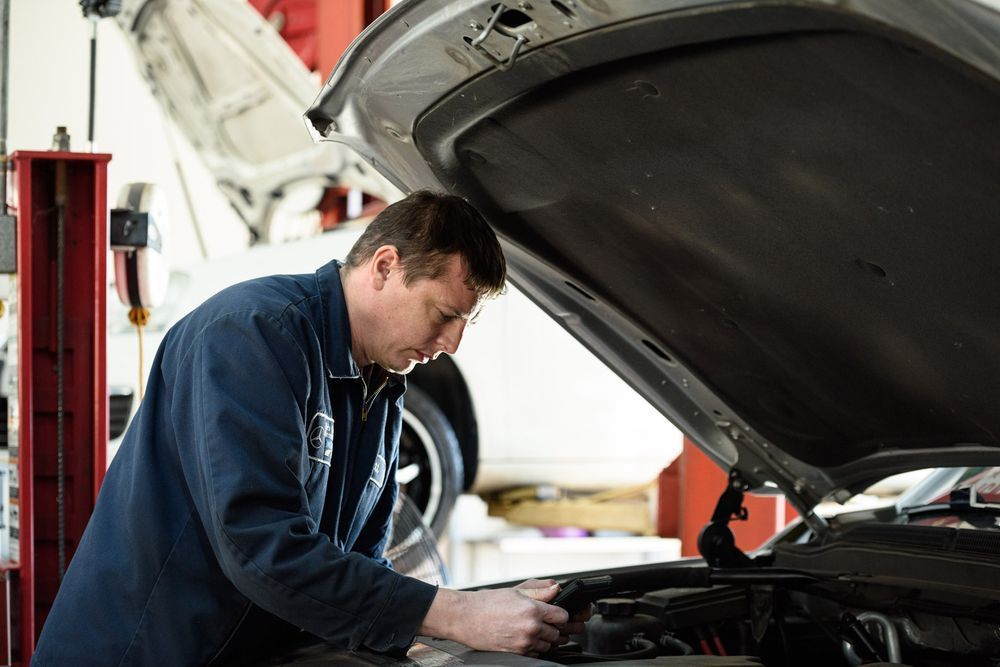 Mechanic inspects car engine in a garage. Wearing blue uniform, hood open.