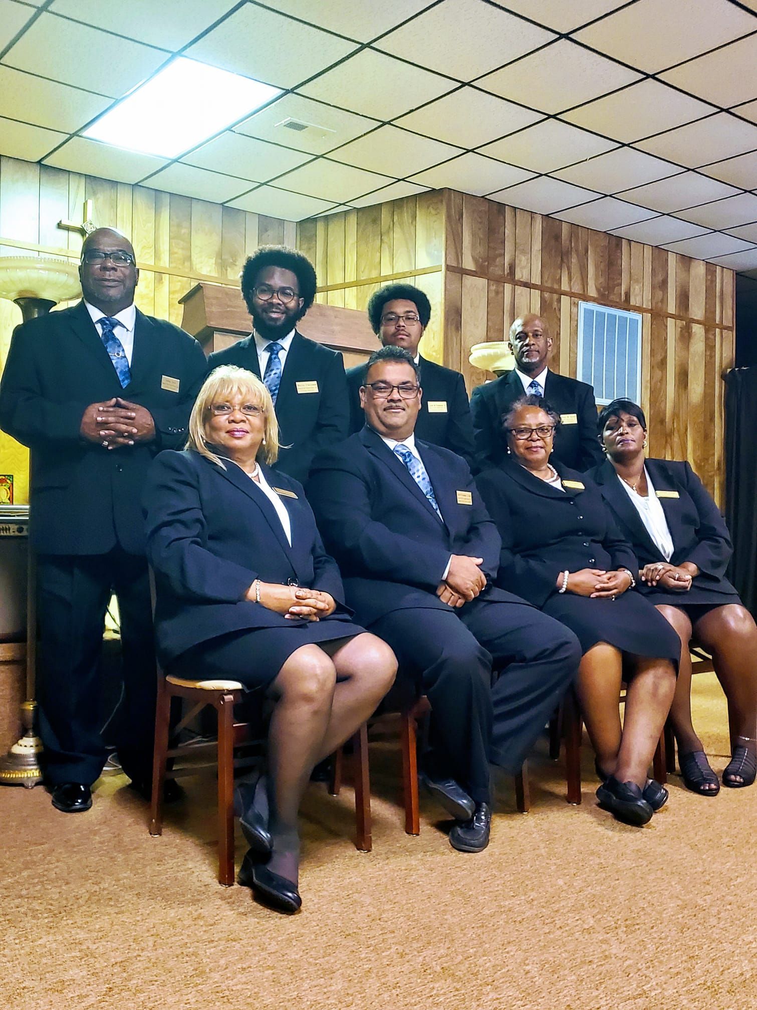 Eight people posed in formal attire in a wood-paneled office, seated and standing in two rows.