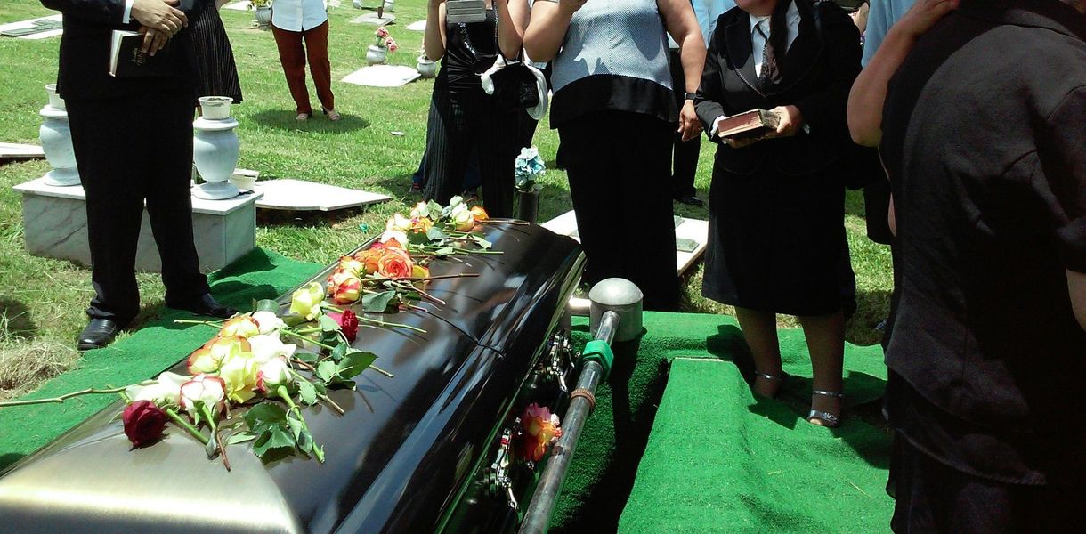 Mourners stand around a flower-covered black coffin on green turf at an outdoor funeral.