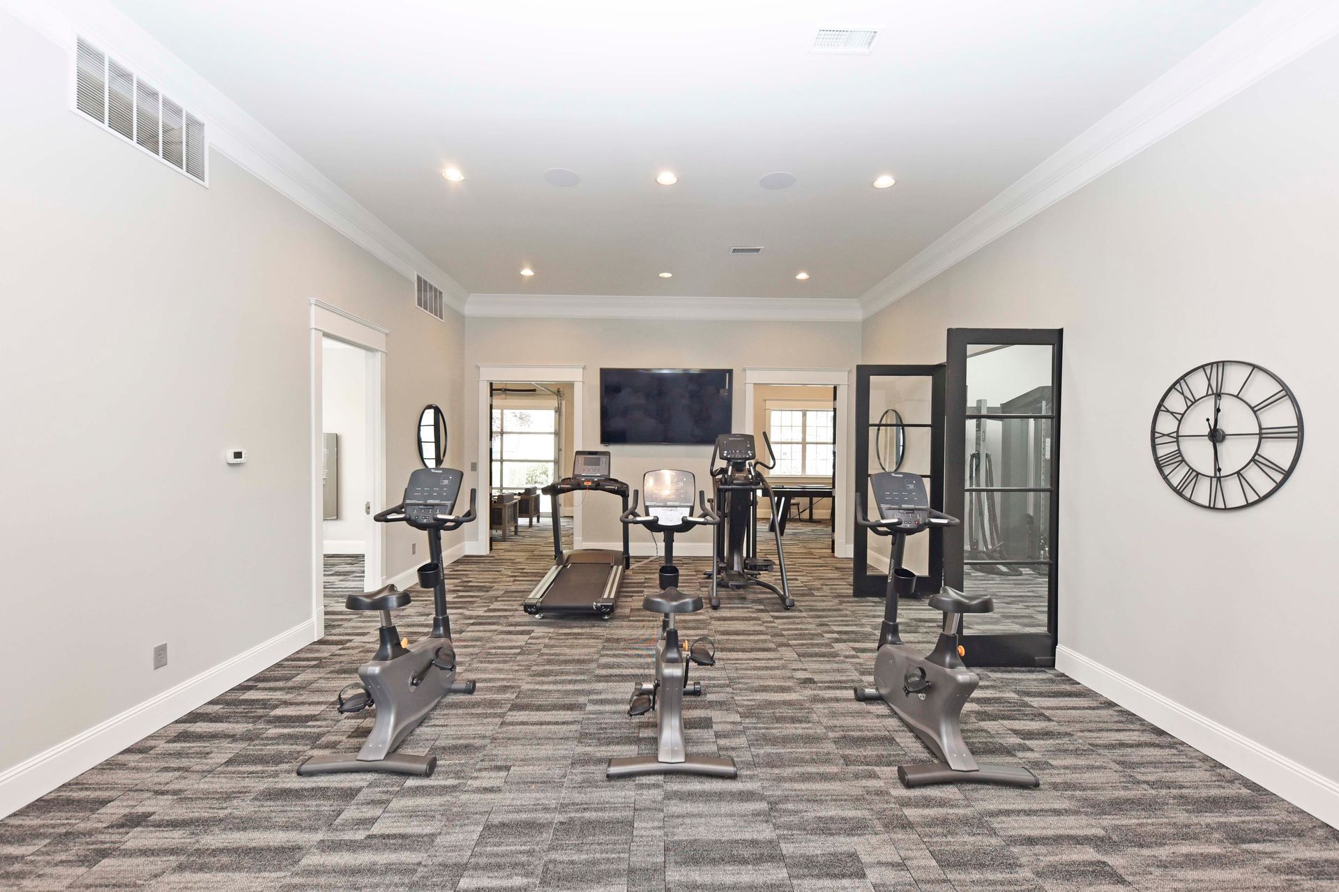 A home gym with exercise bikes, treadmill, and weight machine. Neutral colors with a clock on the wall.