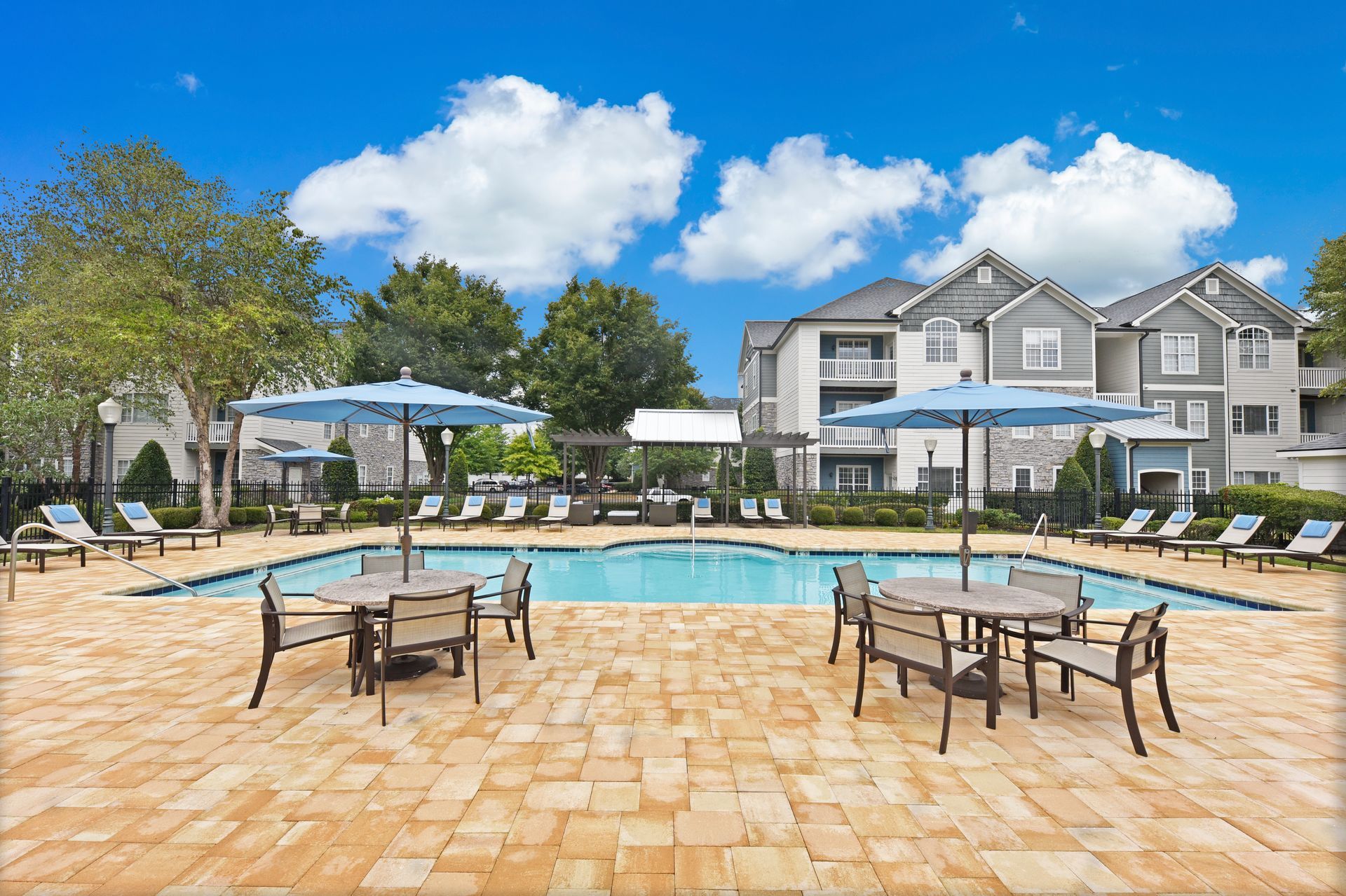 Pool area with lounge chairs, tables, umbrellas, and apartment buildings on a sunny day.