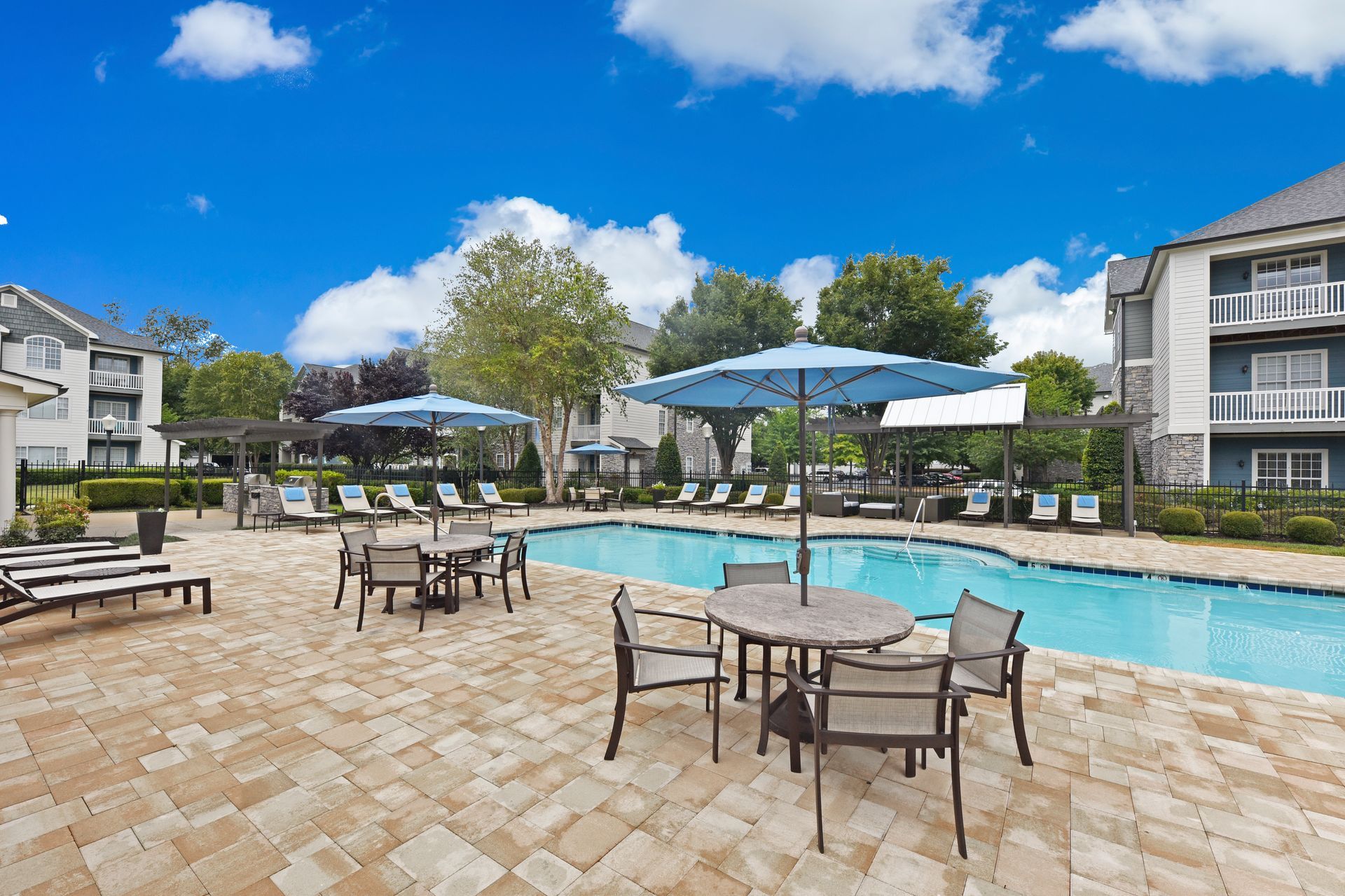 Pool area with tables, chairs, and umbrellas. Apartment buildings in the background, blue sky with clouds.