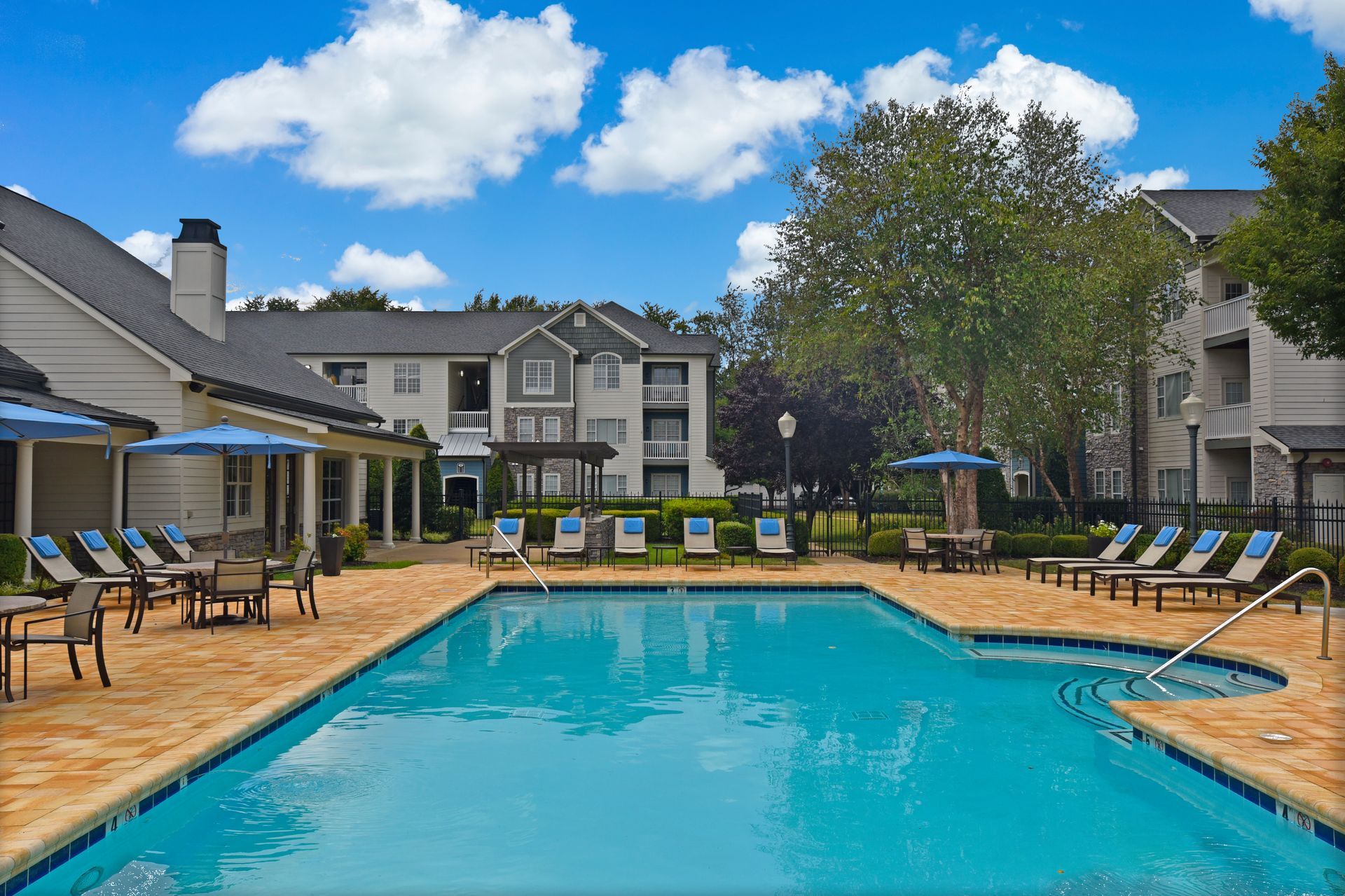 Pool area with lounge chairs, tables, and apartment buildings under a blue sky with clouds.