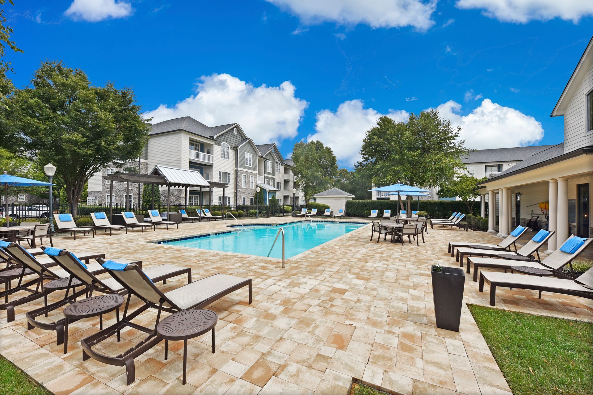Poolside scene with lounge chairs, tables, umbrellas, and apartments under a blue sky.