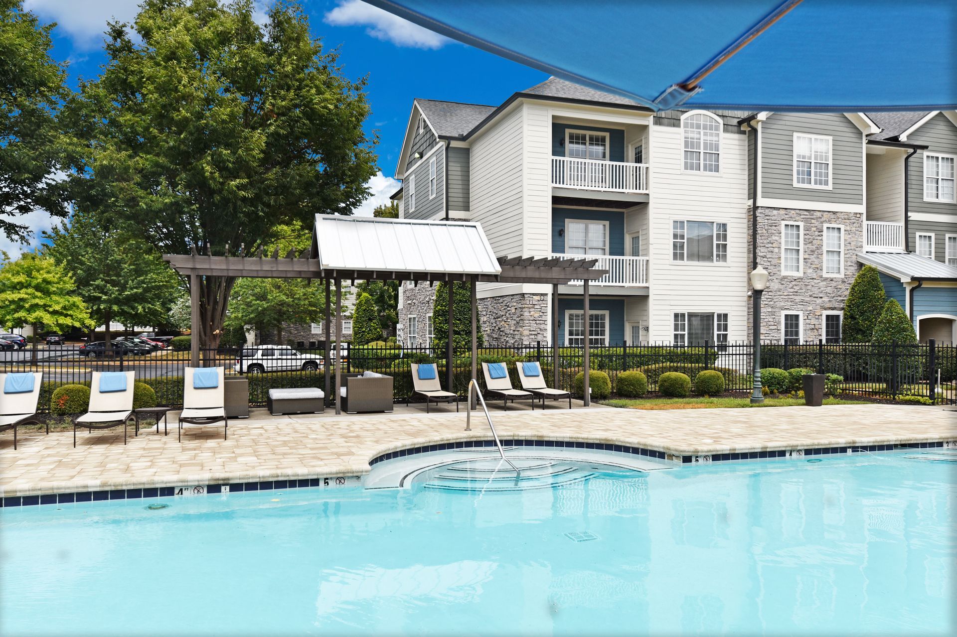 Pool area with lounge chairs, pergola, and apartment building. Blue water, sunny day.