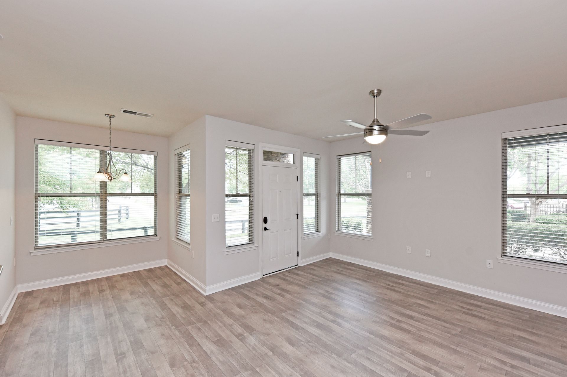 Empty living room with large windows, ceiling fan, and wood-look flooring.