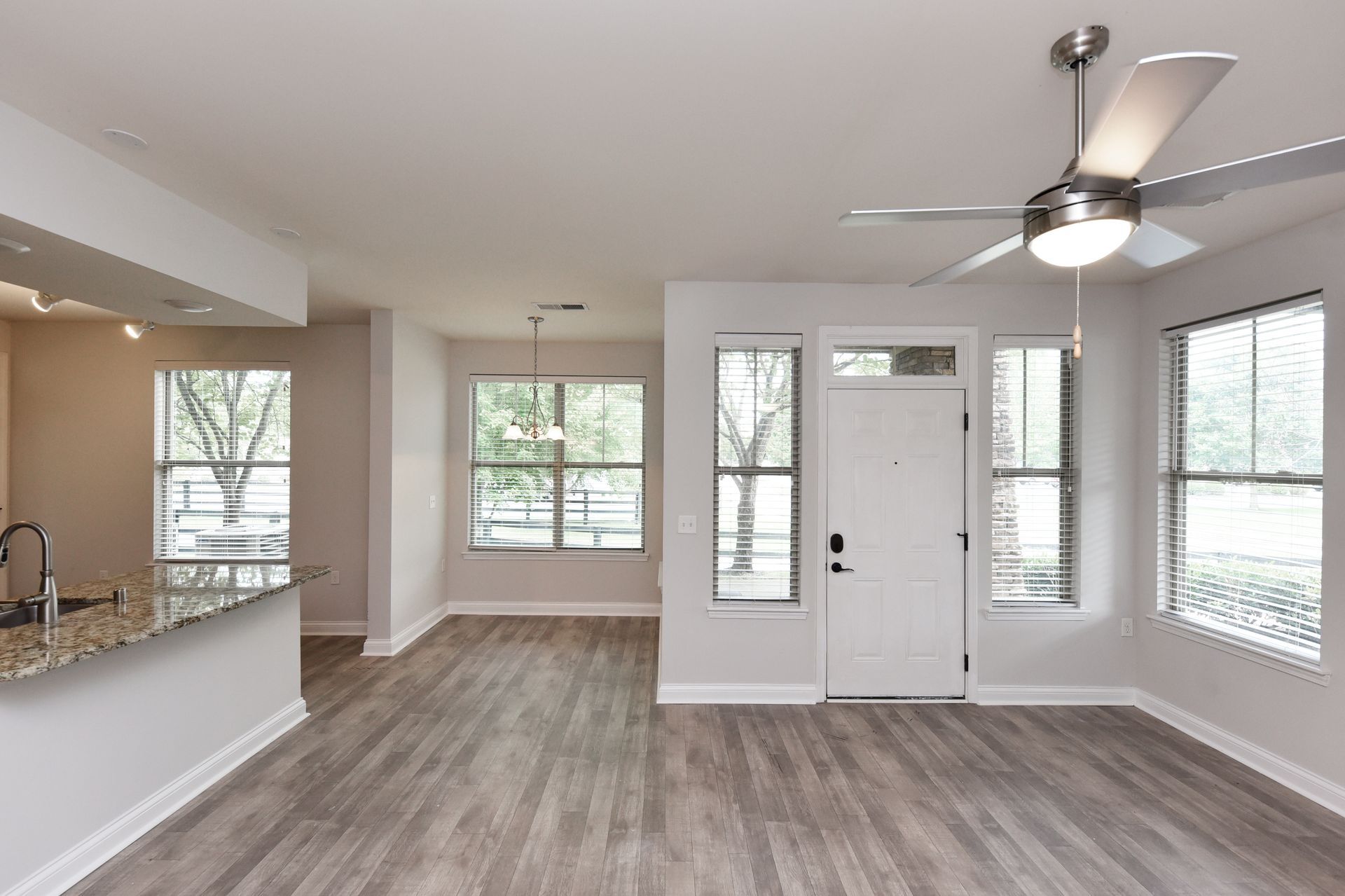 Spacious living area with light gray walls, wood-look floors, windows, and a ceiling fan.