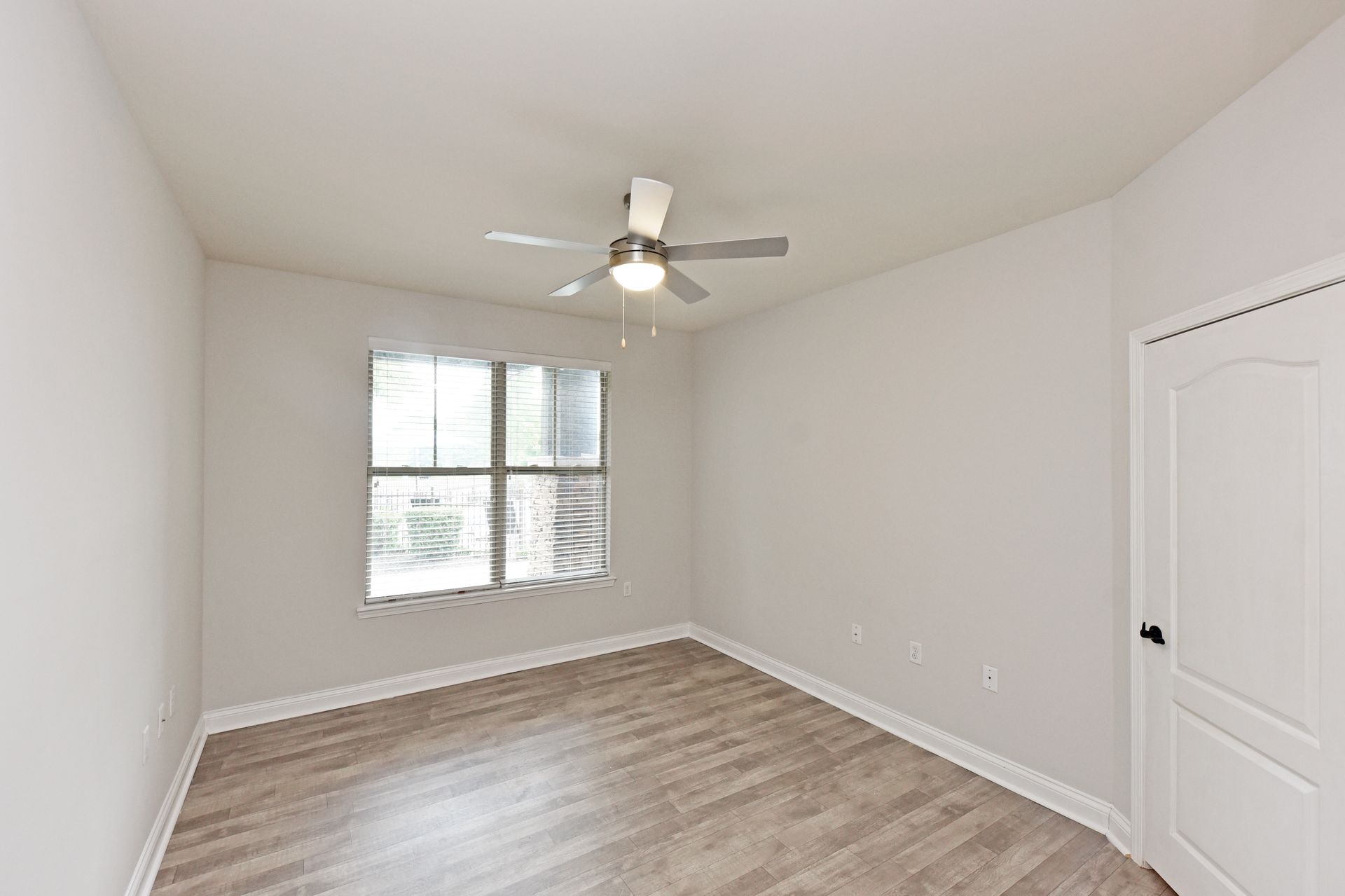 Empty bedroom with a ceiling fan, window, and wood-look flooring. White walls and a white door.