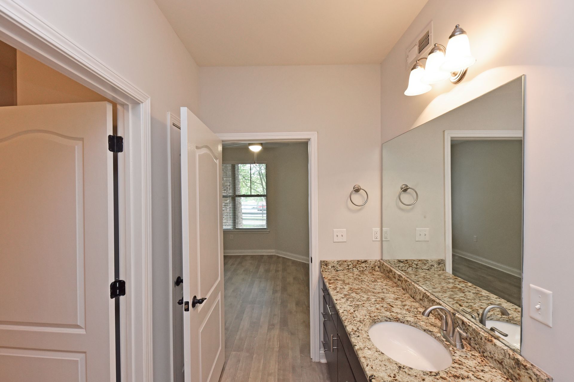Bathroom with granite countertop, mirror, and open doorway to a bedroom with wood flooring.